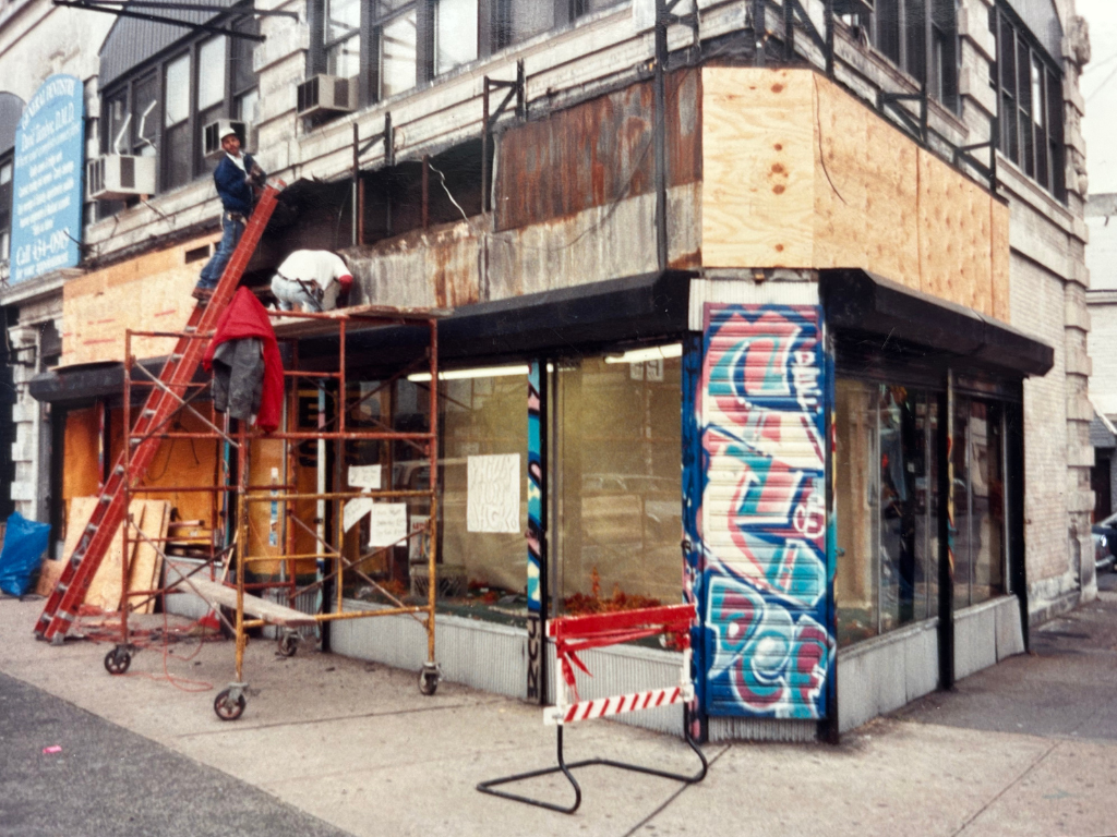 People working on building renovation of a commercial storefront on a city street, with scaffolding, graffiti-covered wall, and plywood panels.