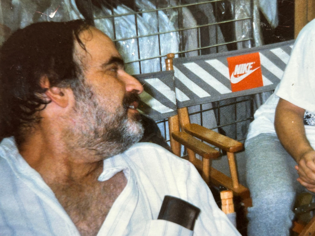 Close-up of a middle-aged man with dark hair and a beard, wearing a white shirt, sitting indoors near a Nike shoe box.