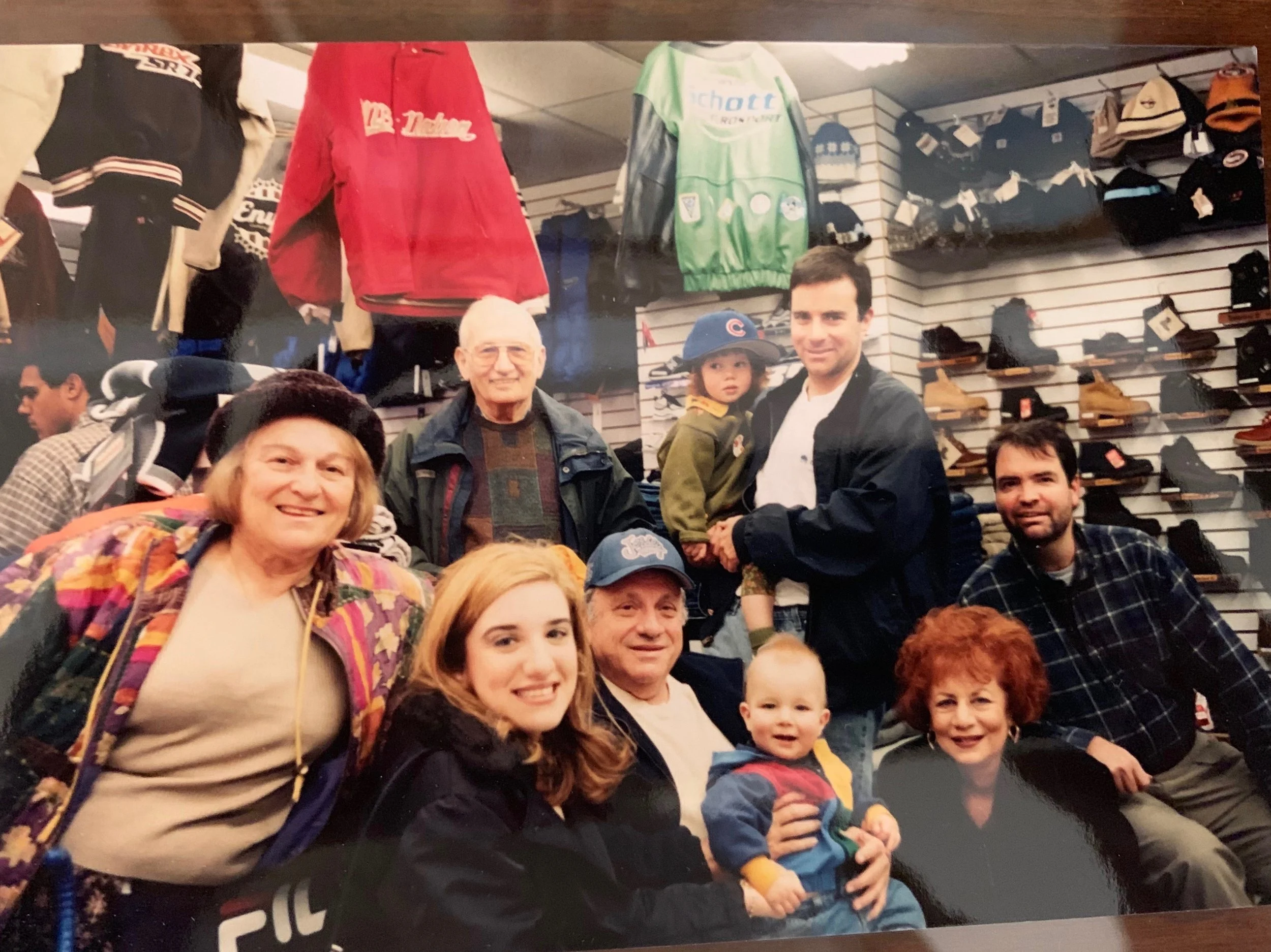 A group of nine people, including children, posed together inside a store that sells outdoor clothing and boots. The background shows various jackets, hats, and shoes on display.