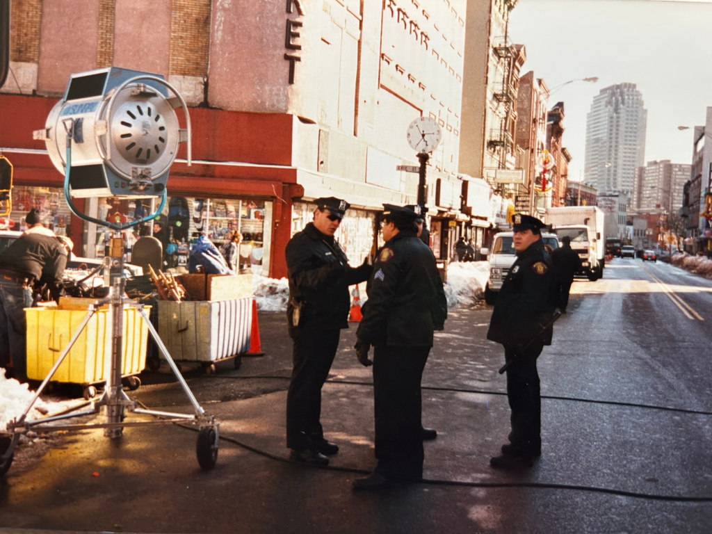 Three police officers stand on a city street in winter, talking near a street vendor with stalls and a large outdoor light, with tall buildings in the background.