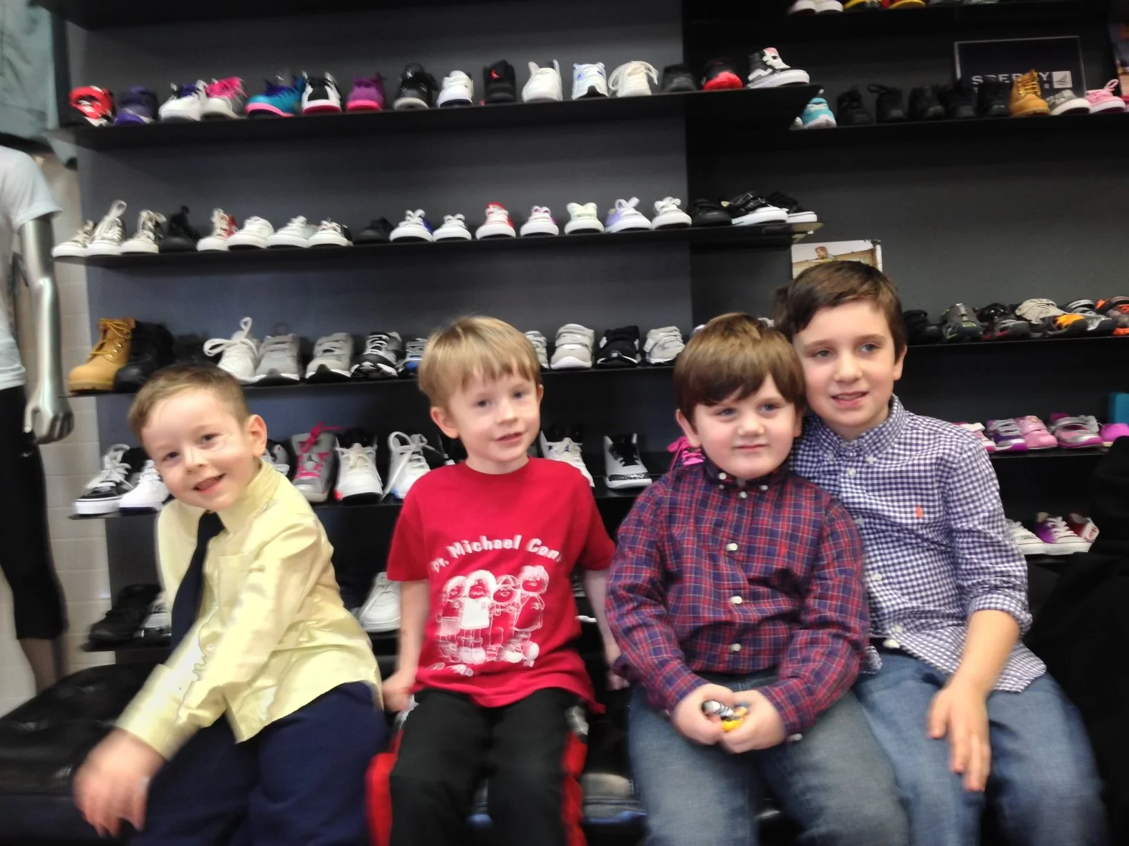 Four young boys sitting in front of a display of various sneakers on shelves, smiling for the photo in a shoe store.