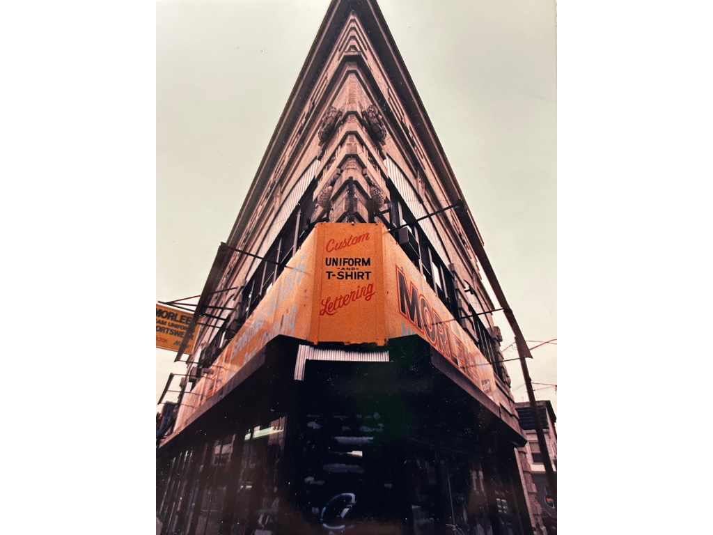 A low-angle view of a corner building adorned with orange and black signage, advertising custom uniforms and lettering services, with multiple stories and a narrow street below.