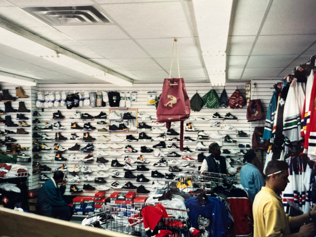 Interior of a shoe and apparel store with various sneakers on white slatwall, clothing racks, and shoppers browsing.