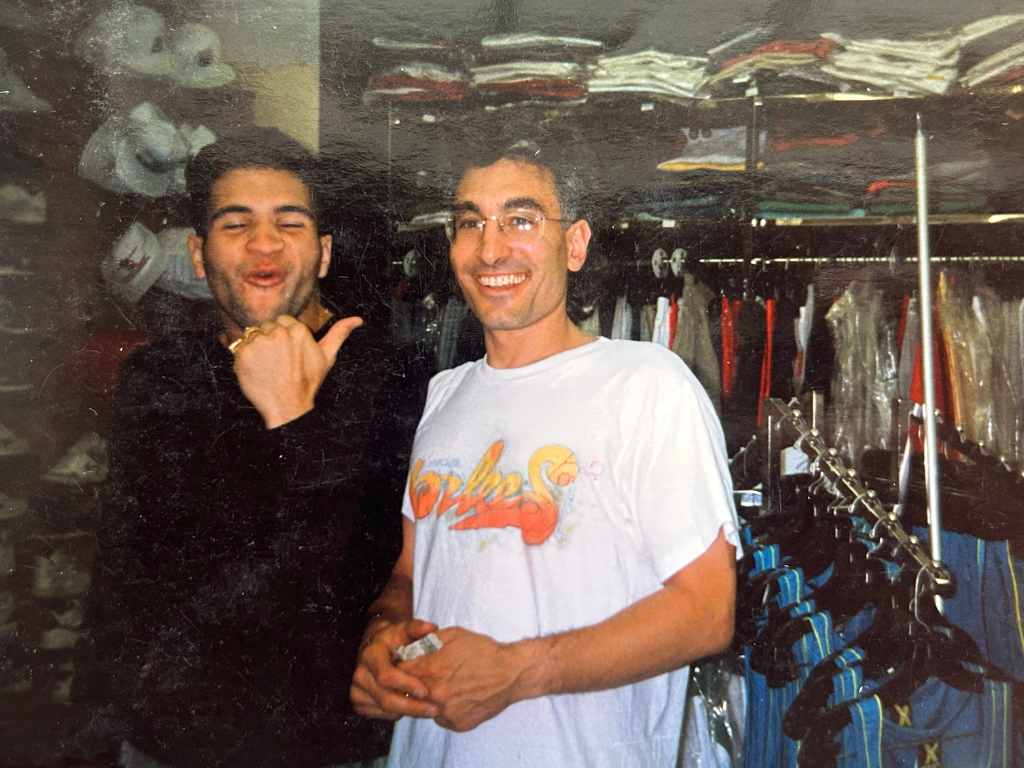 Two men stand inside a clothing store surrounded by racks of clothing and shelves with hats. The man on the left is smiling with his eyes closed, wearing a black jacket, and making a fist gesture. The man on the right is smiling, wearing glasses and