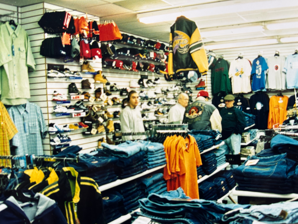 Secondhand clothing store with shelves of jeans, T-shirts, and jackets, and shelves of sneakers and hats, with three customers browsing.