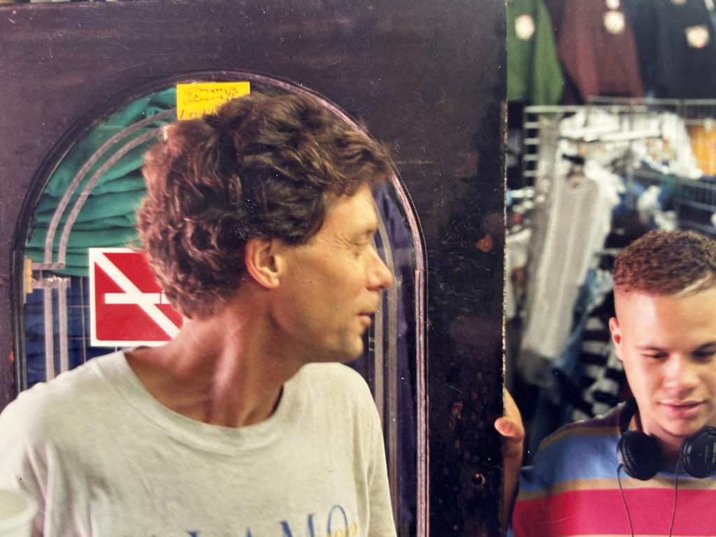 Two young men in a retail store, one with curly hair and wearing headphones, the other with short hair, looking toward the right side of the image. It appears to be a clothing or thrift store with shelves of clothing in the background.
