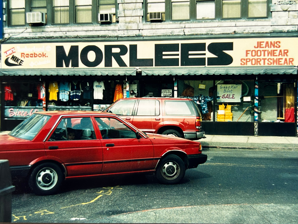 Street view showing a storefront named MORLEES selling jeans, footwear, and sportswear, with signs for a sneaker boot sale, and two red cars parked in front.