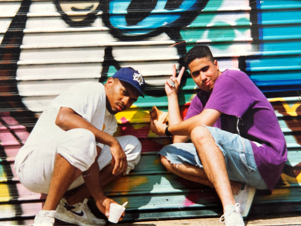 Two young men sitting in front of a colorful graffiti wall. One wears a white outfit and a blue cap, while the other is in a purple shirt and jeans, making a peace sign.