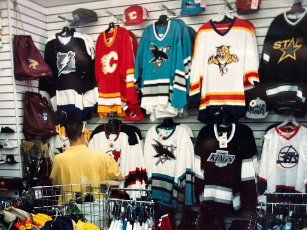 A person shopping in a sports apparel store with hockey jerseys displayed on racks and wall, featuring teams like San Jose Sharks, Calgary Flames, Texas Stars, Florida Panthers, and Dallas Stars.