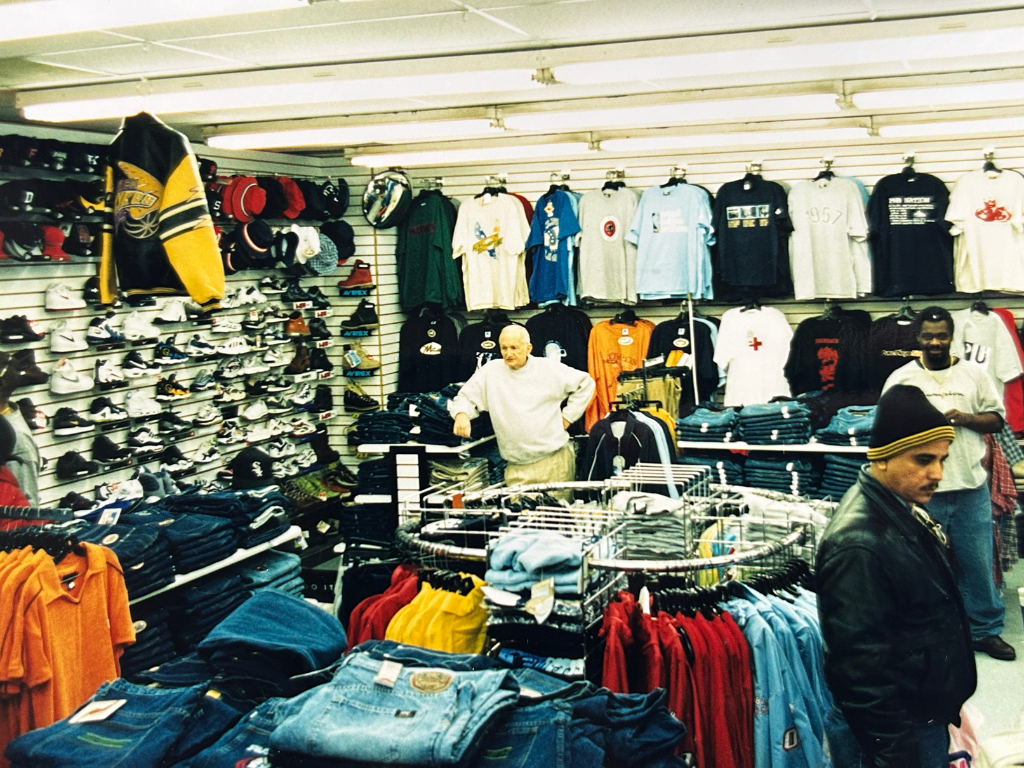 Inside a clothing store with racks of shirts, pants, hats, and sneakers, and three men browsing merchandise.
