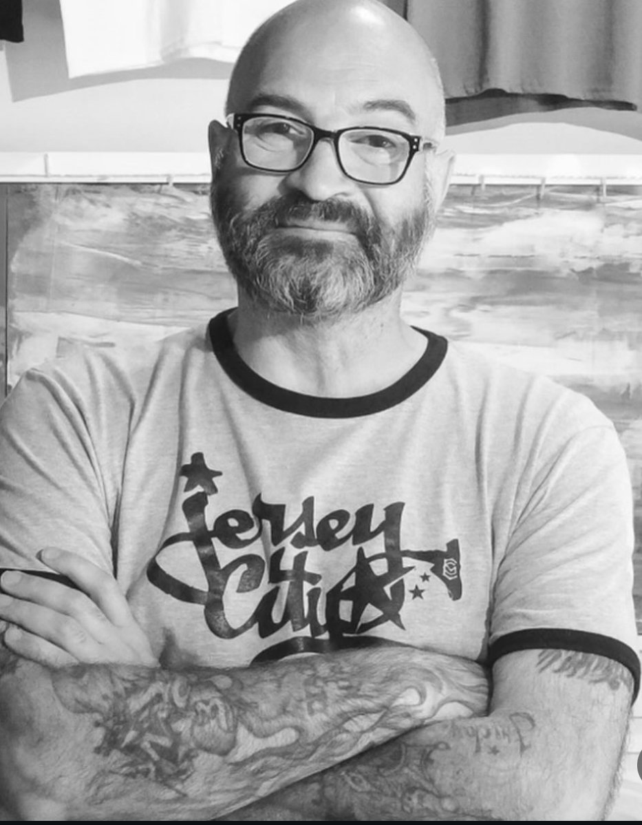 JUAN CHEESE VASQUEZ, Creative director of Morlees. Black and white photo of a smiling man with glasses, a beard, and tattoos, wearing a t-shirt with a graphic design and crossed arms.