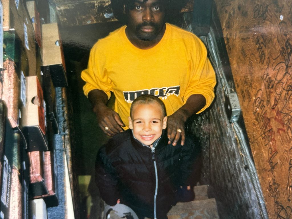 A man and a young boy smiling inside a narrow, cluttered space that appears to be a storage area or small room. The man is wearing a yellow sweatshirt, and the boy is wearing a black jacket. They are looking up at the camera, which is positioned abov