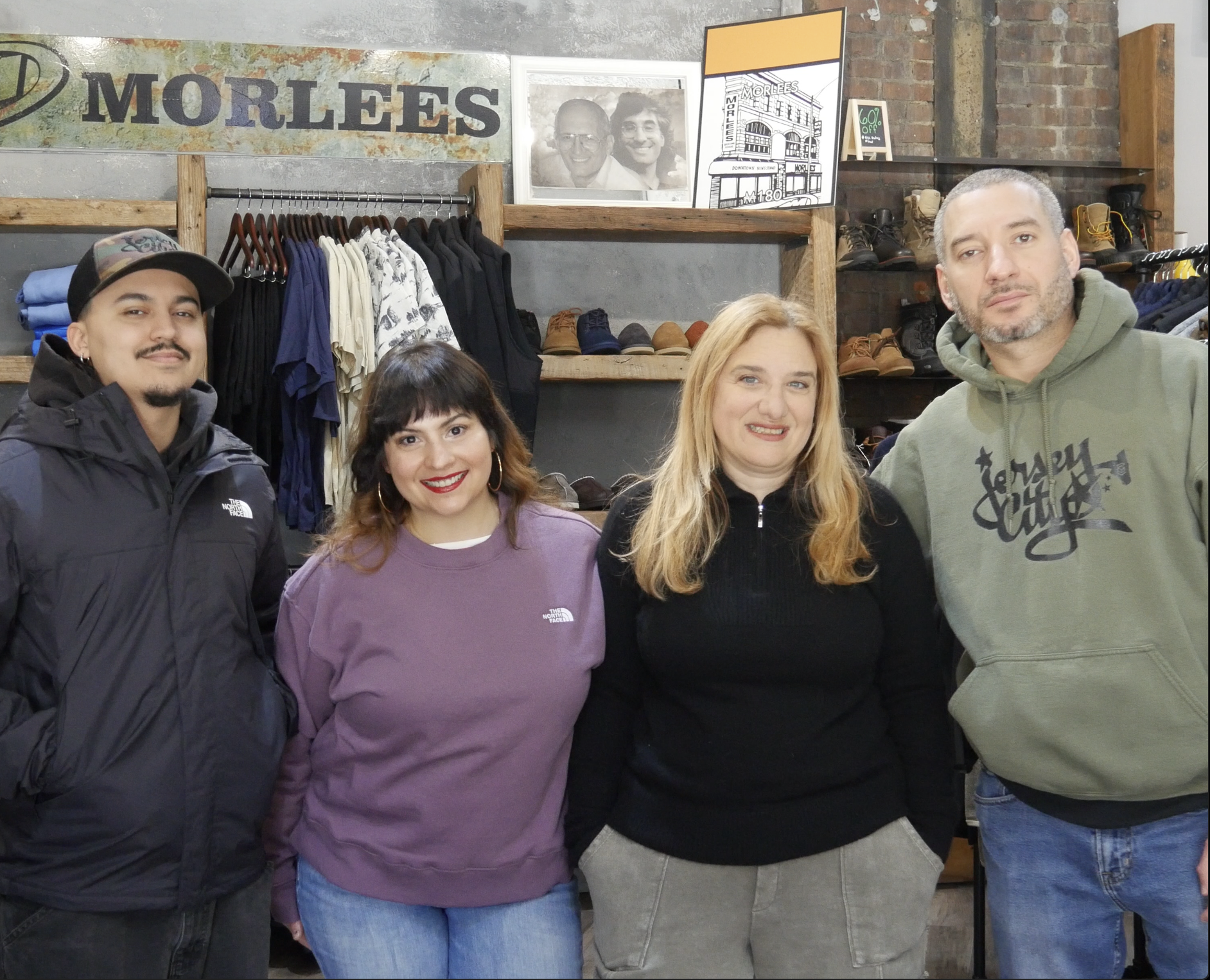 Group of four people standing inside a clothing store, smiling at the camera. Behind them are shelves with shoes and hanging shirts, and a banner with the word 'MORLEES' on the wall.
