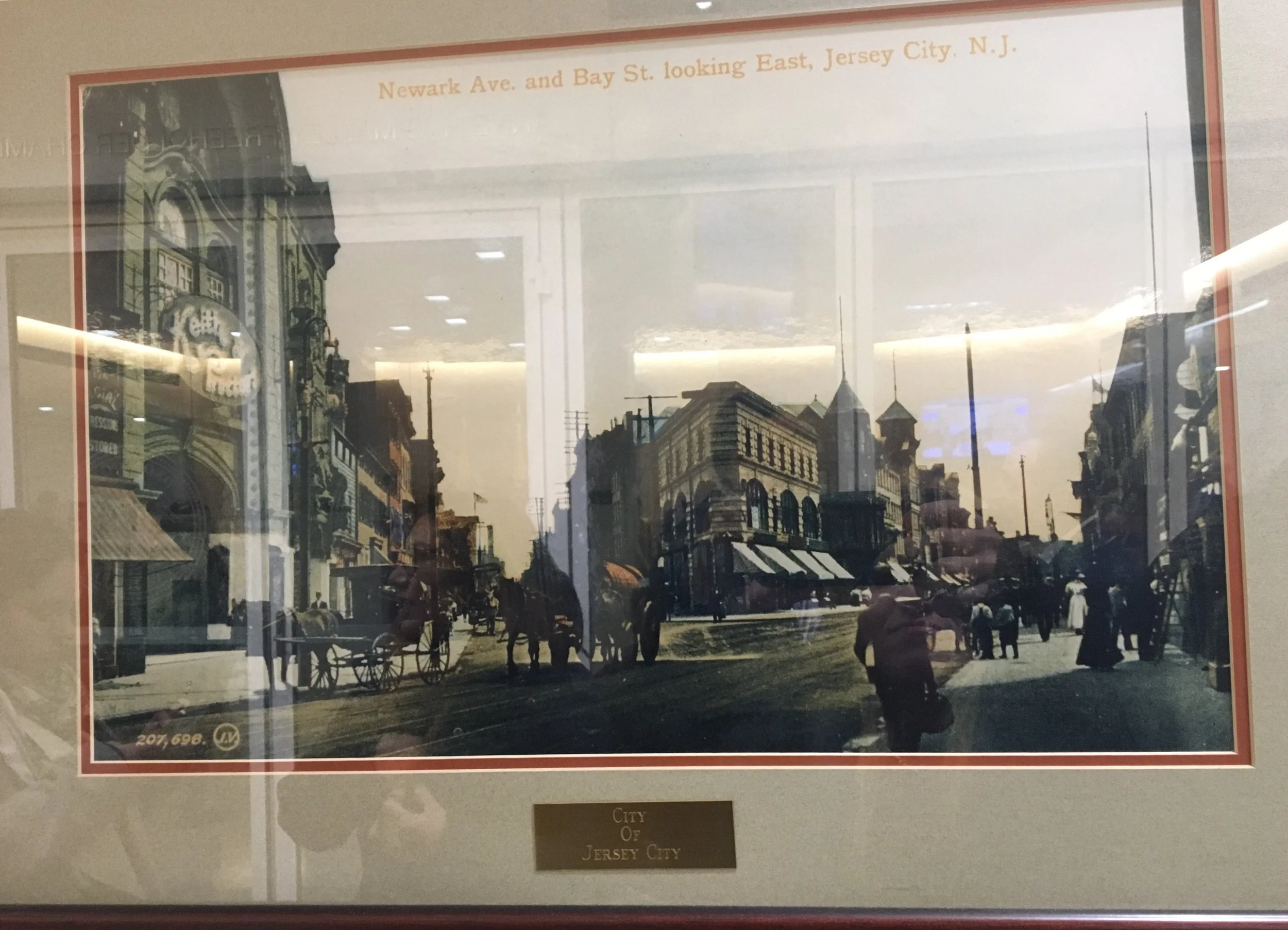 Historical black and white photograph of a street scene in Jersey City, showing old buildings with storefronts, horse-drawn carriages, and pedestrians, titled "Newark Ave. and Bay St. looking East, Jersey City, N.J."