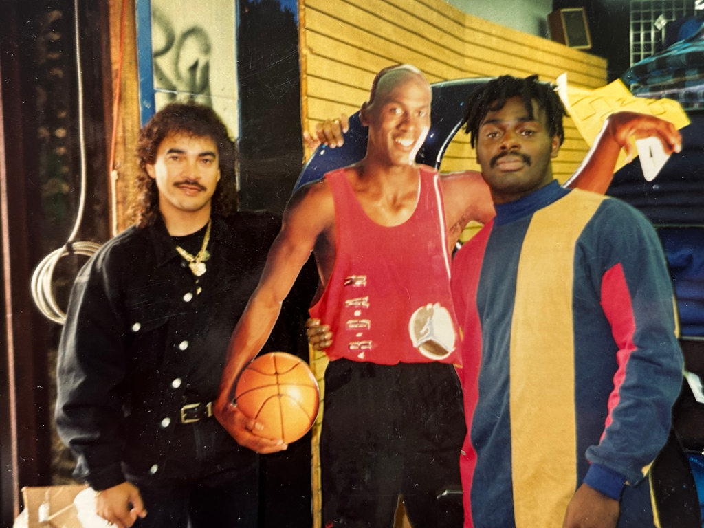 Three men posing indoors, with the middle person holding a basketball, and the person on the right has dreadlocks and is wearing a multicolored jacket.
