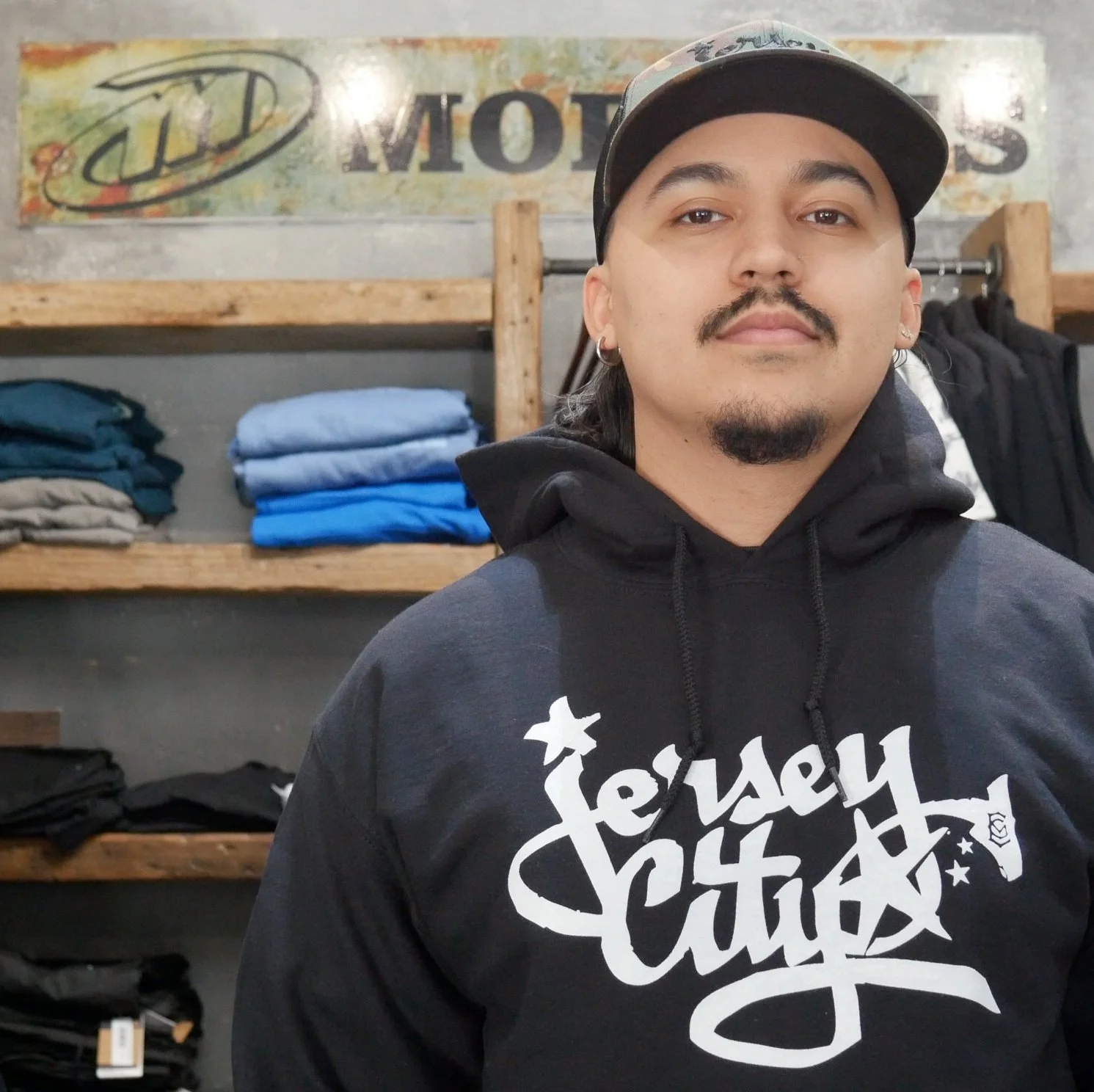 Angel , sales representative of Morlees, Jersey City, NJ. Description: A person with long dark hair, mustache, and goatee wearing a black hoodie and a cap standing inside a clothing store.