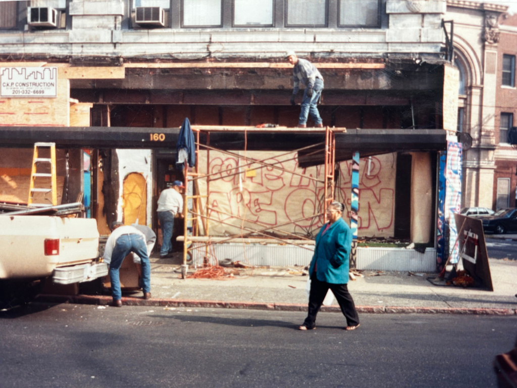 Construction workers working on a building facade with graffiti, with a woman walking on the sidewalk in front.
