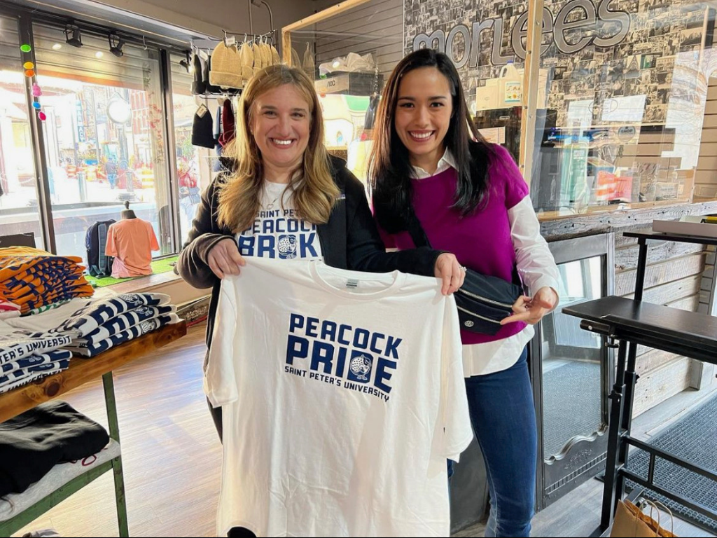 Two women smiling inside a store, holding a white T-shirt with 'PEACOCK PRIDE SAINT PETER'S UNIVERSITY' printed on it, surrounded by stacks of T-shirts and merchandise.