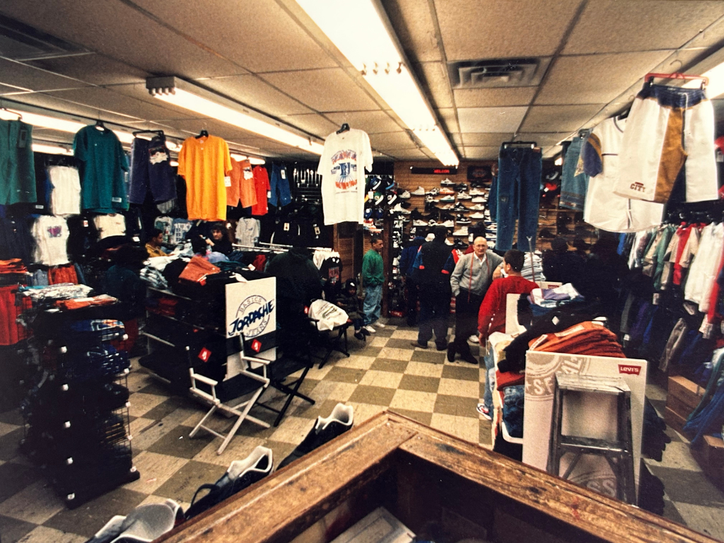 Inside a clothing store with racks of colorful shirts and pants, and several customers shopping and browsing.
