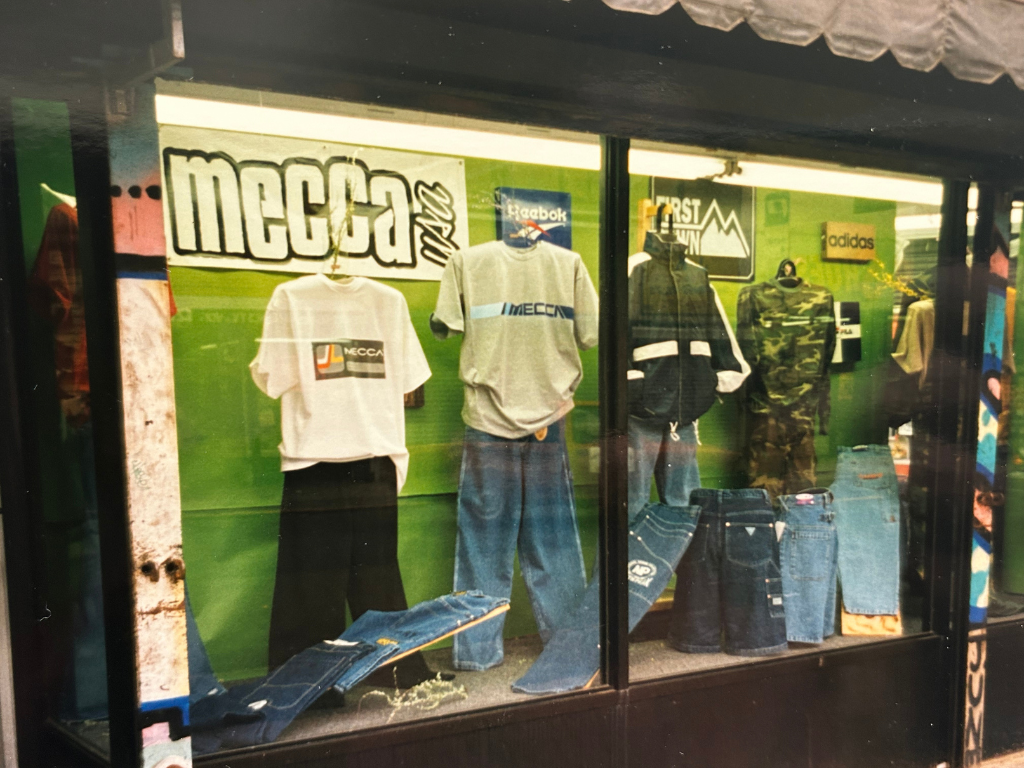 A storefront display showcasing casual clothing including t-shirts, jeans, sweatshirts, and camouflage apparel, with various brand logos and signage in the background.