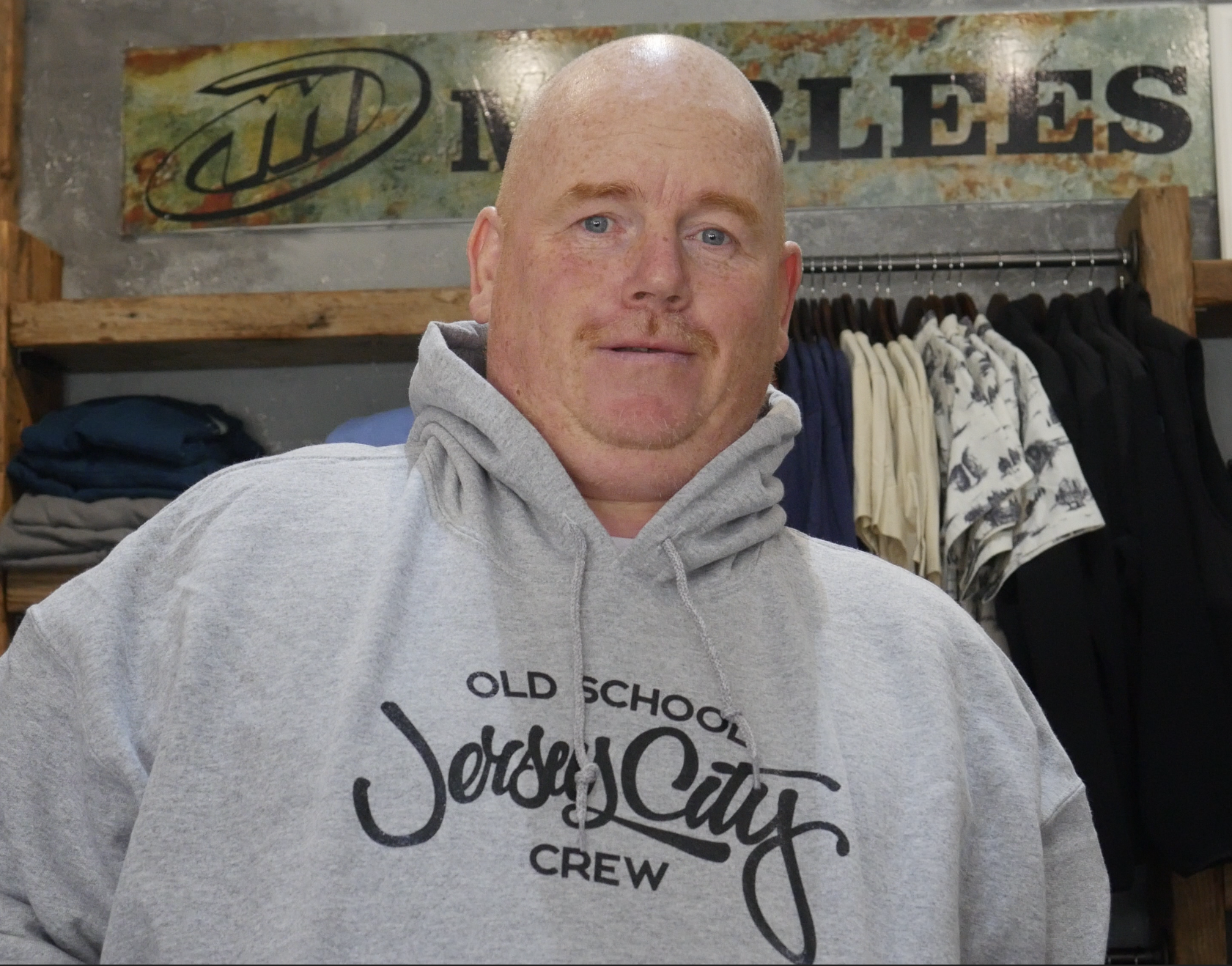 Earl , sales representative of Morlees, Jersey City, NJ. Description:A bald man with a ginger beard wearing a gray hoodie that says 'Old School Jersey City Crew' standing inside a store with clothing racks in the background.