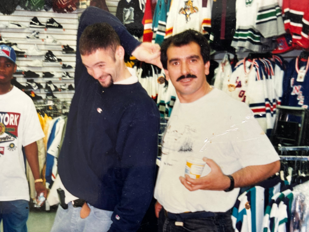 Two men posing in a sports apparel store, one smiling and the other holding a cup, surrounded by jerseys and sneakers.