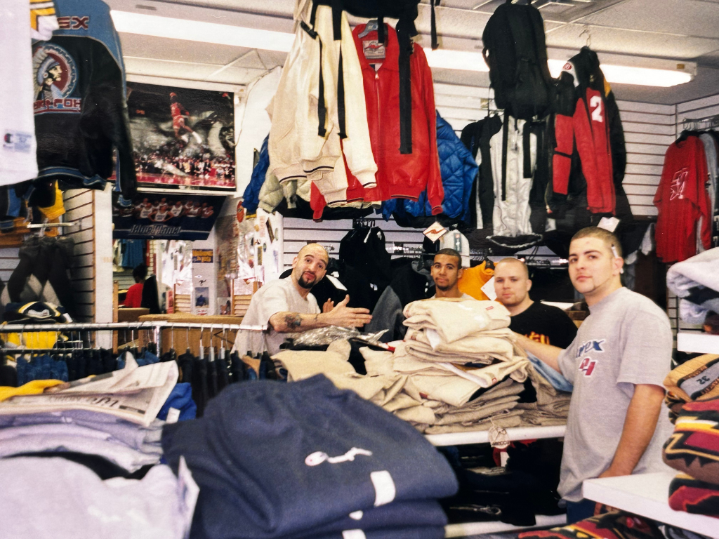 Three men working at a clothing store checkout counter with stacks of folded pants, jackets hanging above, and various clothing items surrounding them.