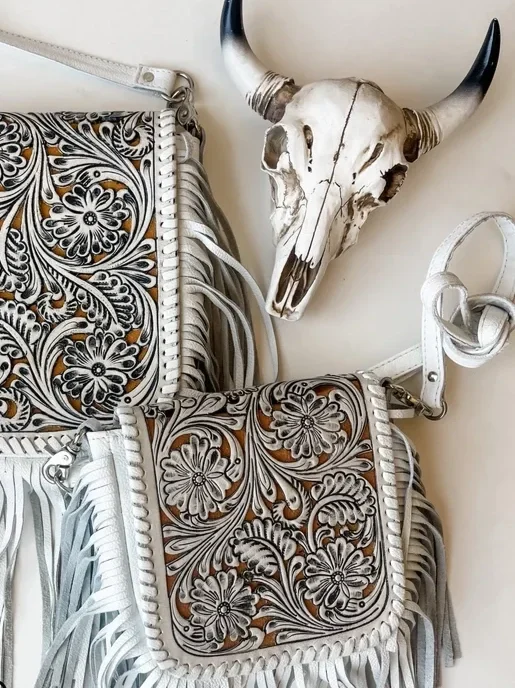A white bull skull with black-tipped horns hangs on a wall, next to two intricately patterned handbags with floral designs and fringe details.