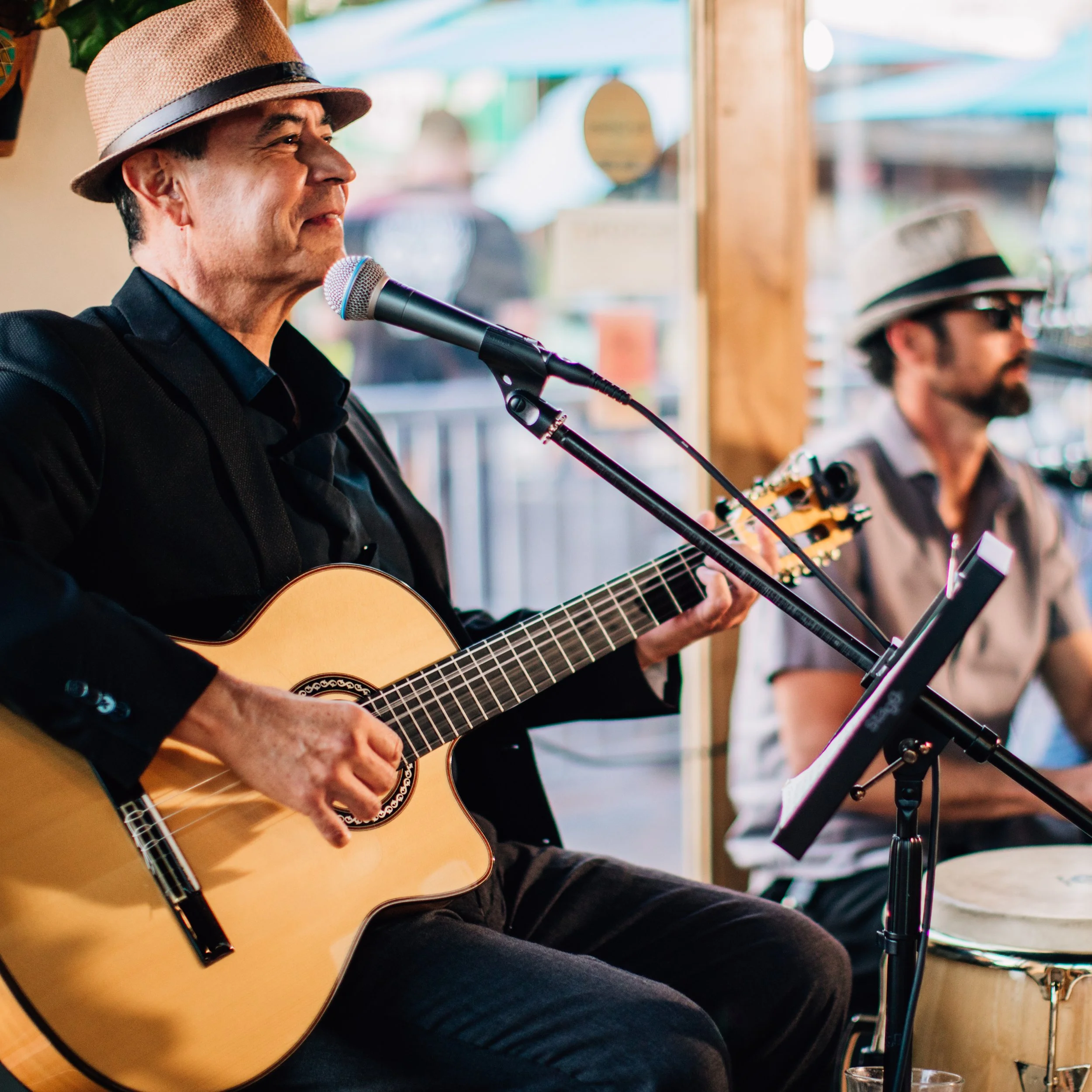 Two musicians performing at 89Agave Cantina; one playing guitar and singing into a microphone, the other playing percussion in the background.