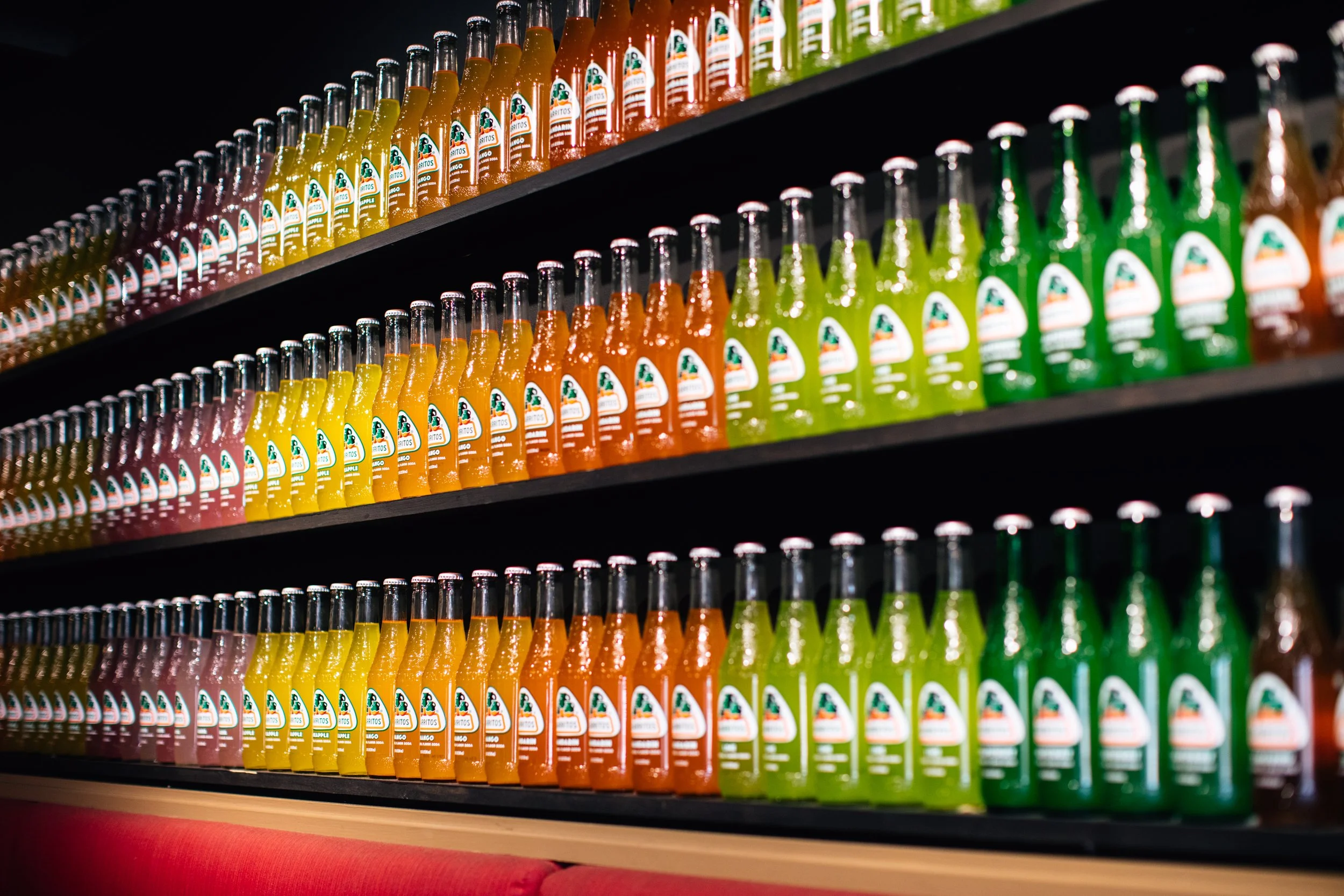 Shelves filled with rows of colorful glass bottles of Jarritos soda, arranged by color, in a store setting.