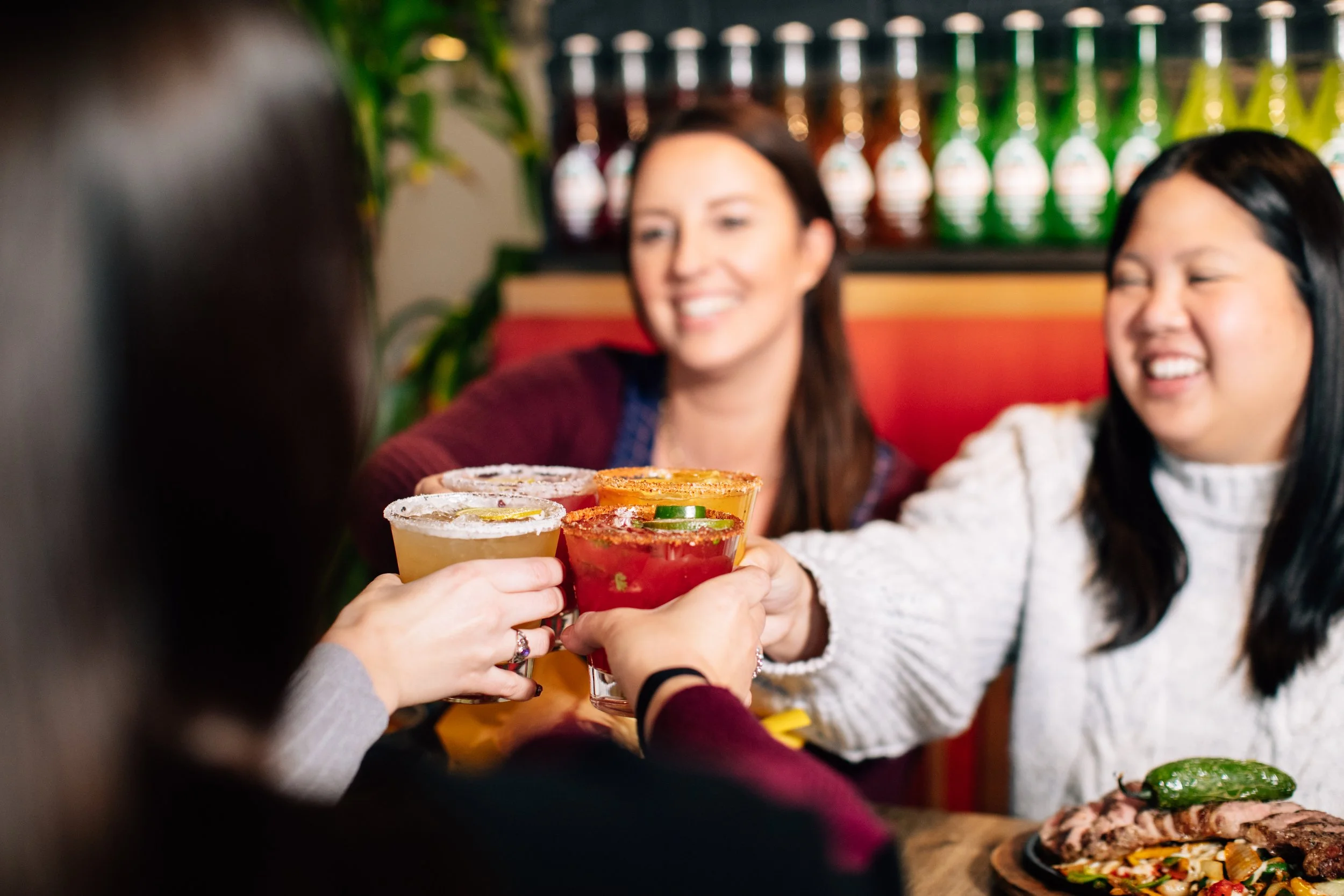 Four women clinking glasses of cocktails at a restaurant table, smiling and enjoying each other's company.