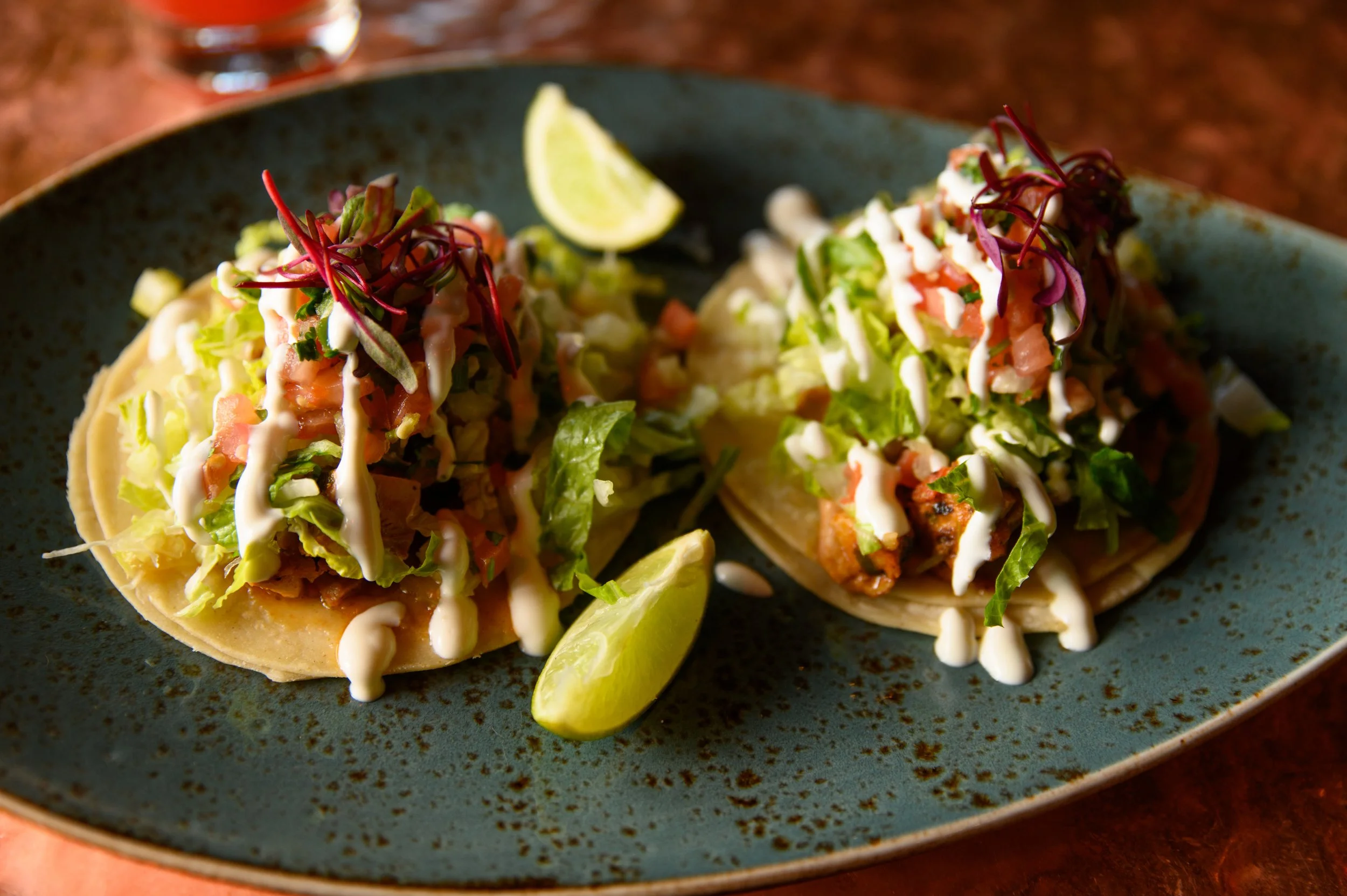 Two tacos filled with lettuce, diced tomatoes, and shredded cheese, topped with a drizzle of white sauce, served with lime wedges on a blue oval plate.