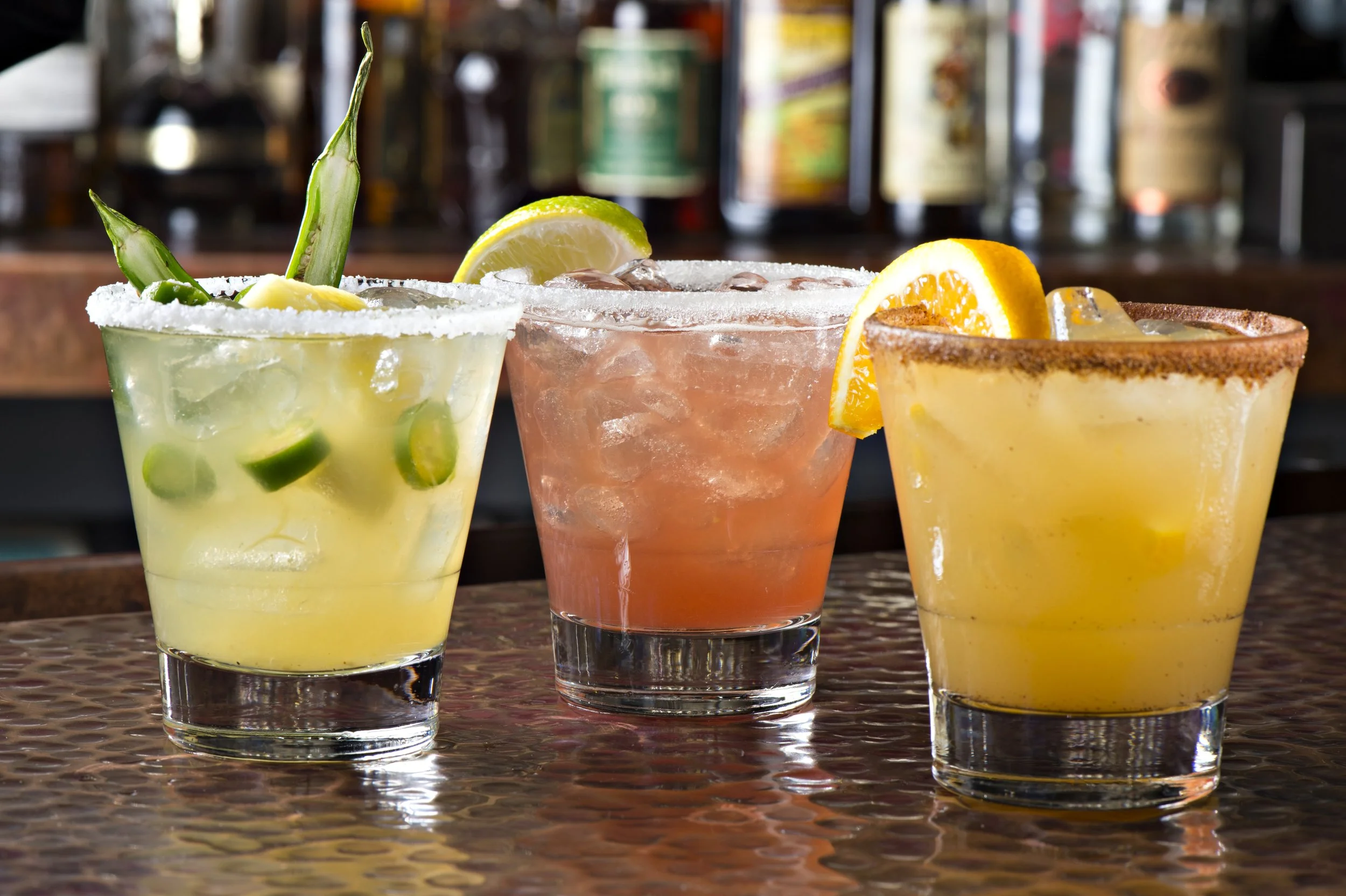 Three colorful cocktails with ice and garnishes on a textured bar surface, with a blurred bar background.