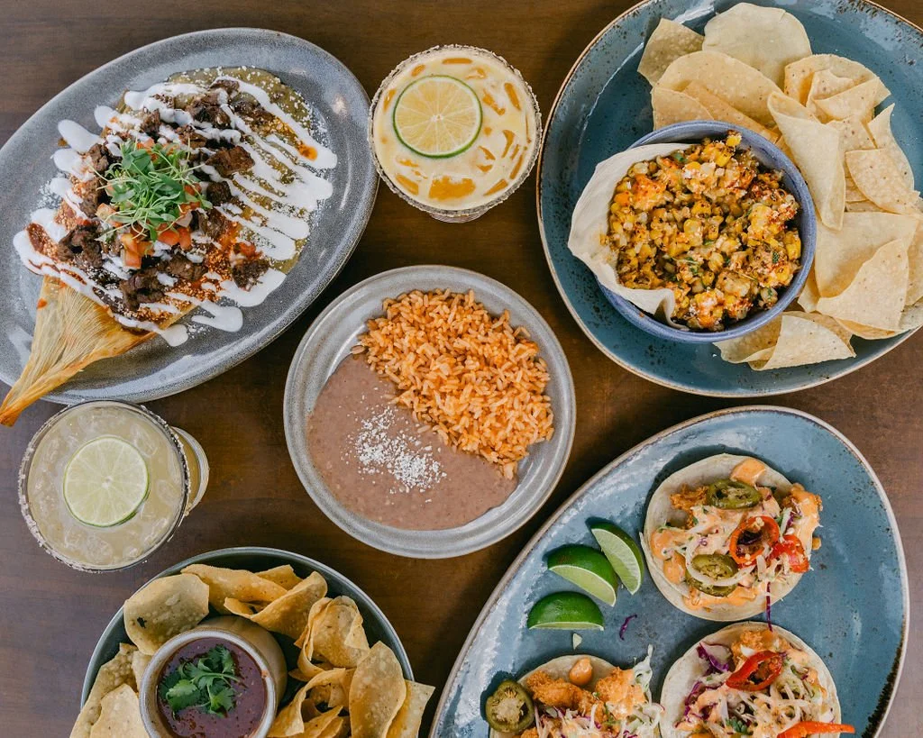 Assorted Mexican dishes served on blue and gray plates, including tacos, rice, beans, nachos, and drinks with lime slices, on a wooden table.