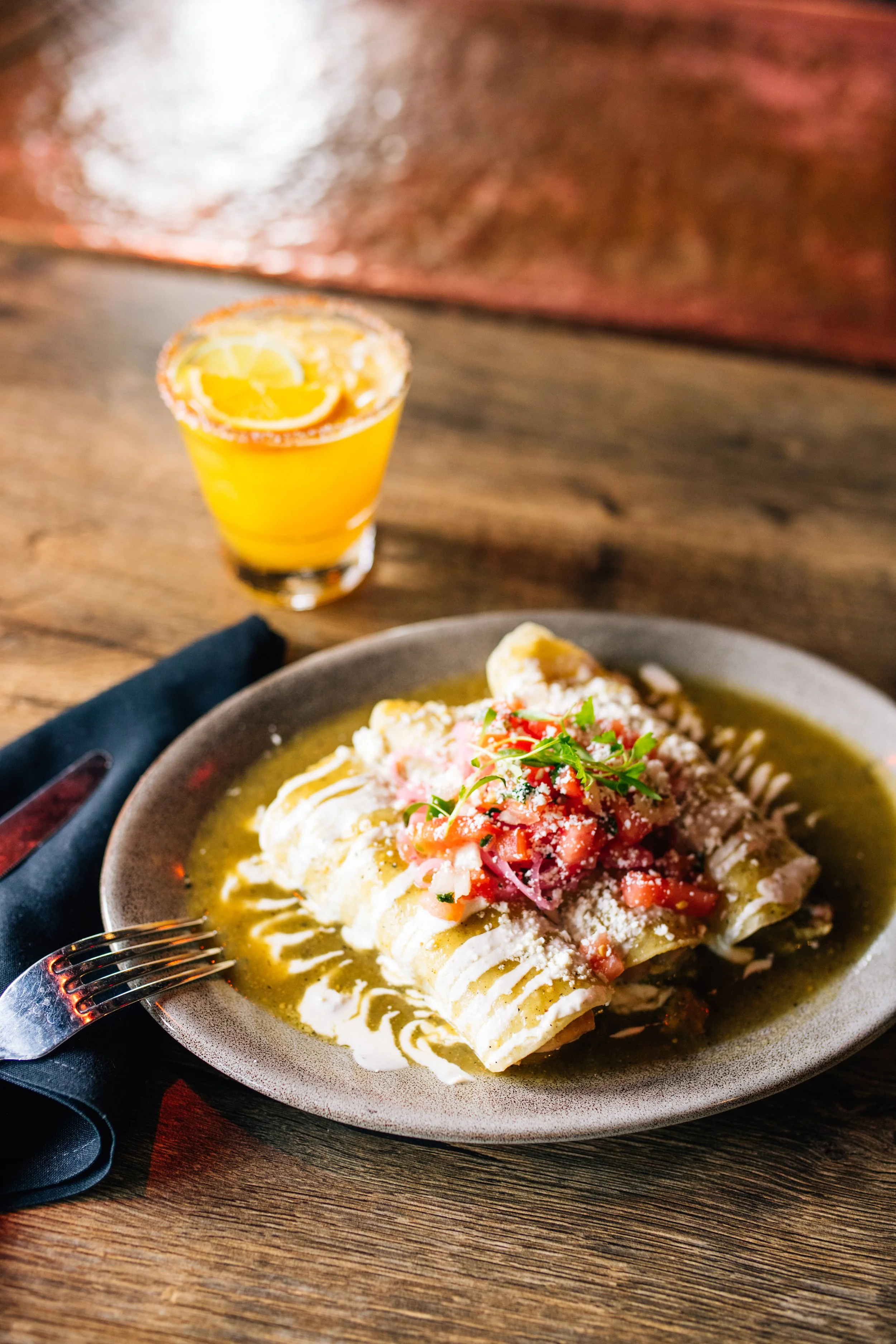 A plate of enchiladas topped with tomato, onion, and cilantro, garnished with cheese, with a side of sauce, served on a rustic wooden table. A glass of orange beverage with lemon slices is in the background.