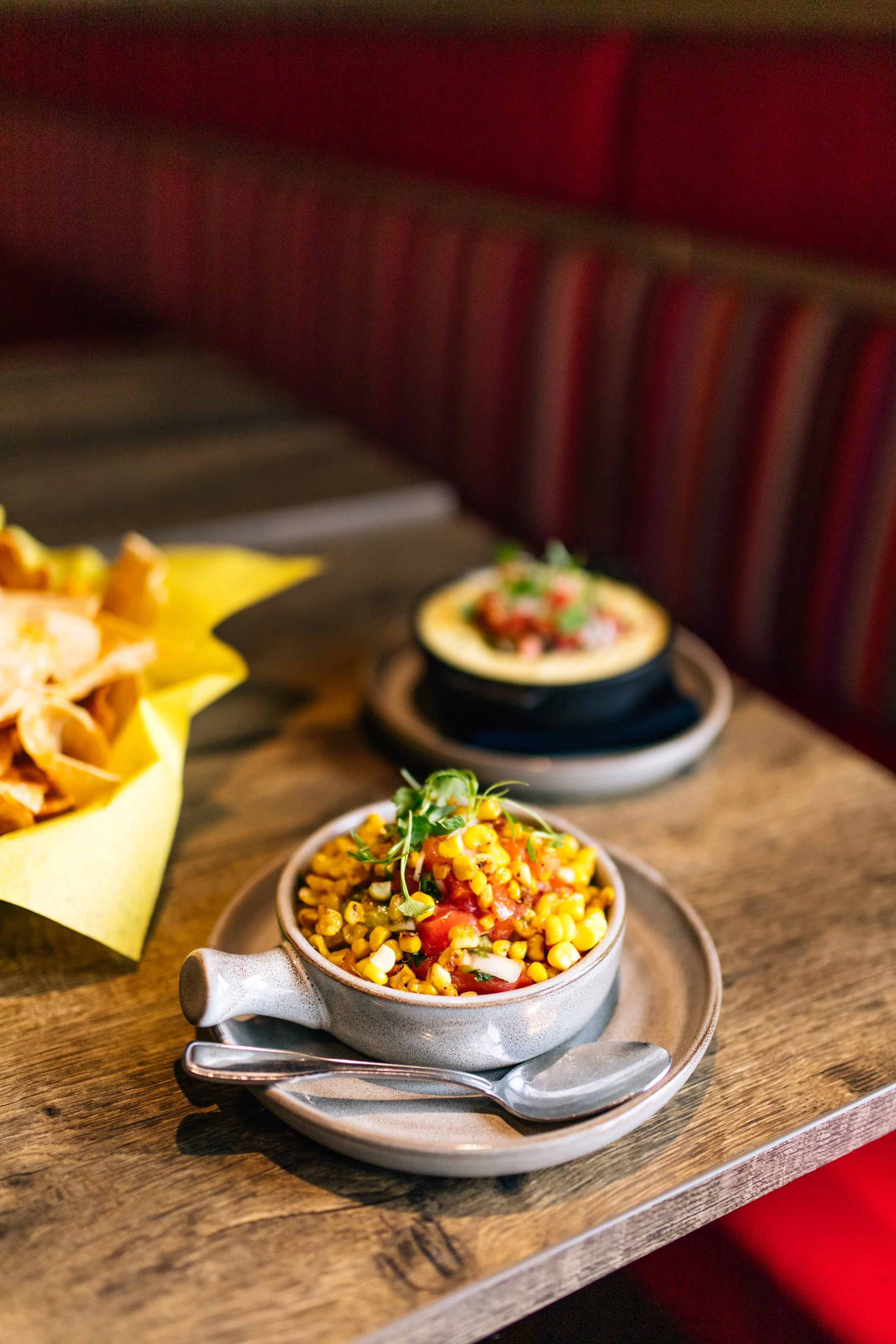 A bowl of corn salad garnished with greens, placed on a wooden table along with a spoon. In the background, there is a small dish with a layered dip or spread. To the left, there is a basket of tortilla chips wrapped in yellow paper.