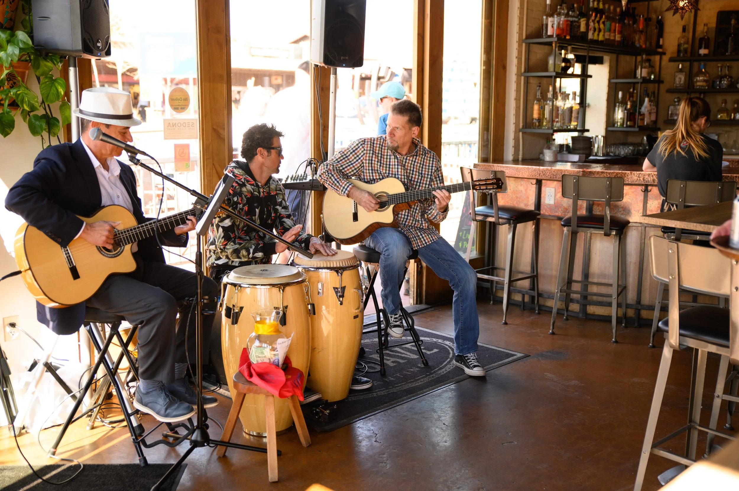 A band of three musicians performing inside at 89Agave Cantina. They are playing guitars and percussion instruments, with a bar and shelves of liquor in the background.