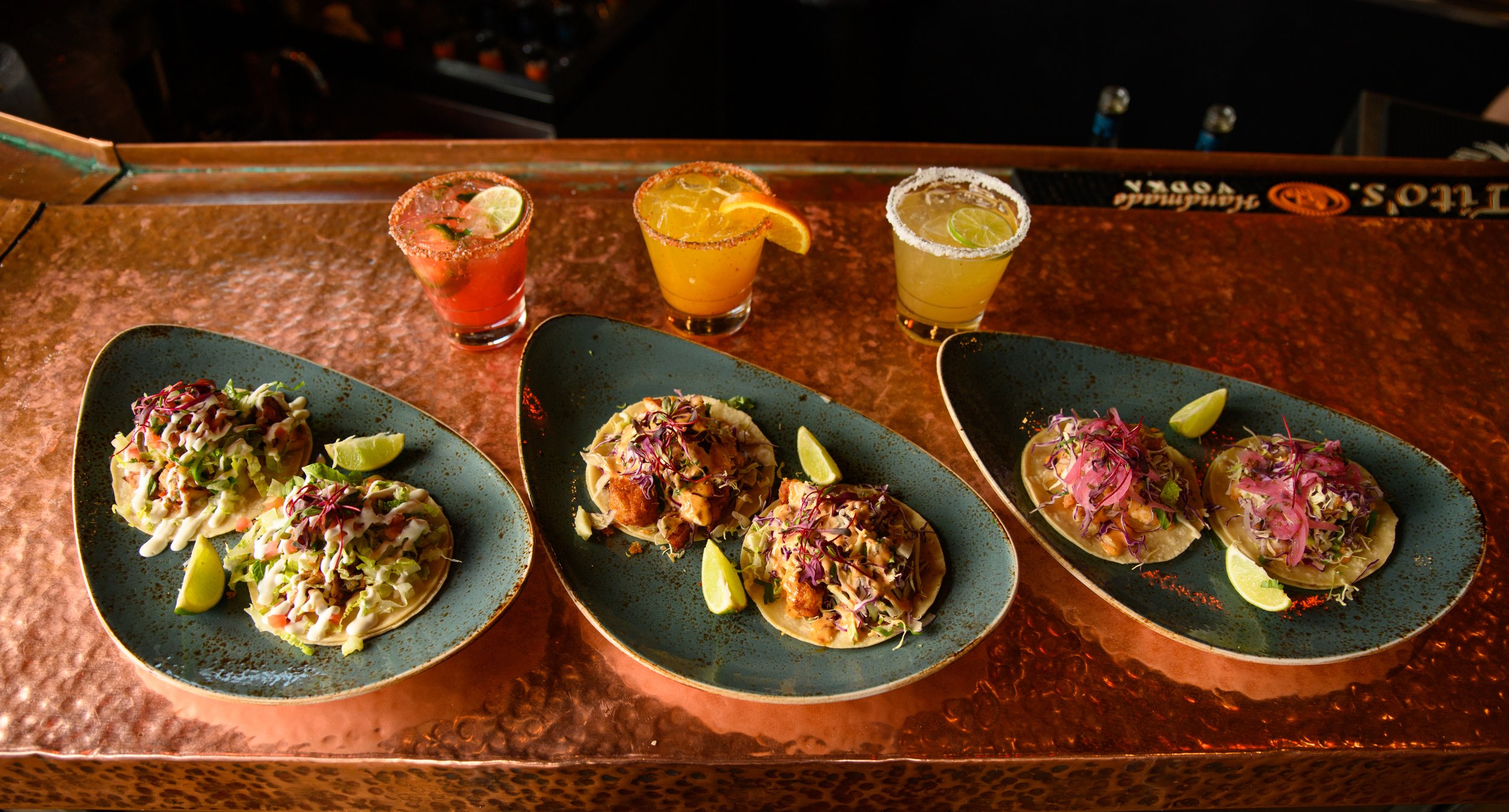 Three plates of tacos with garnishes and three cocktails on a bar counter.