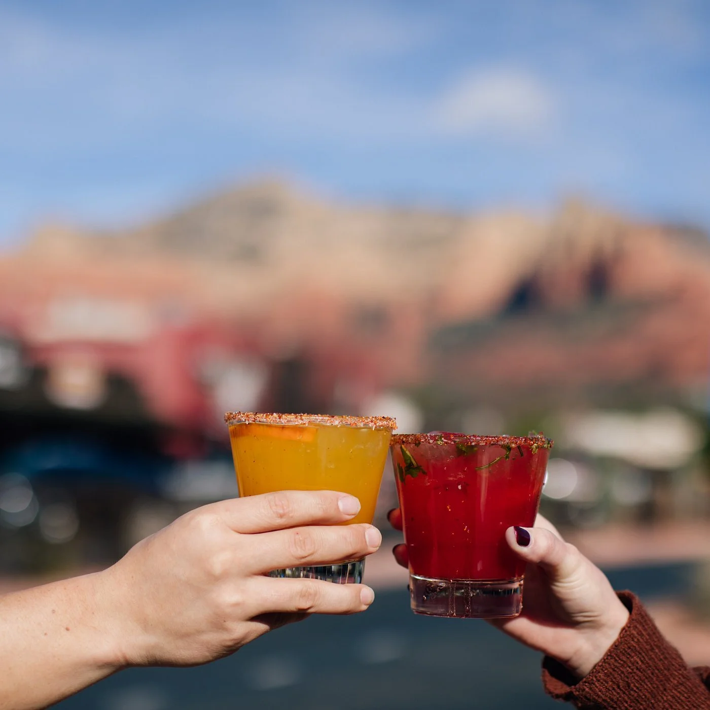 Two hands holding glasses of colorful drinks with salted rims, outdoors with blurred mountains and buildings in the background.