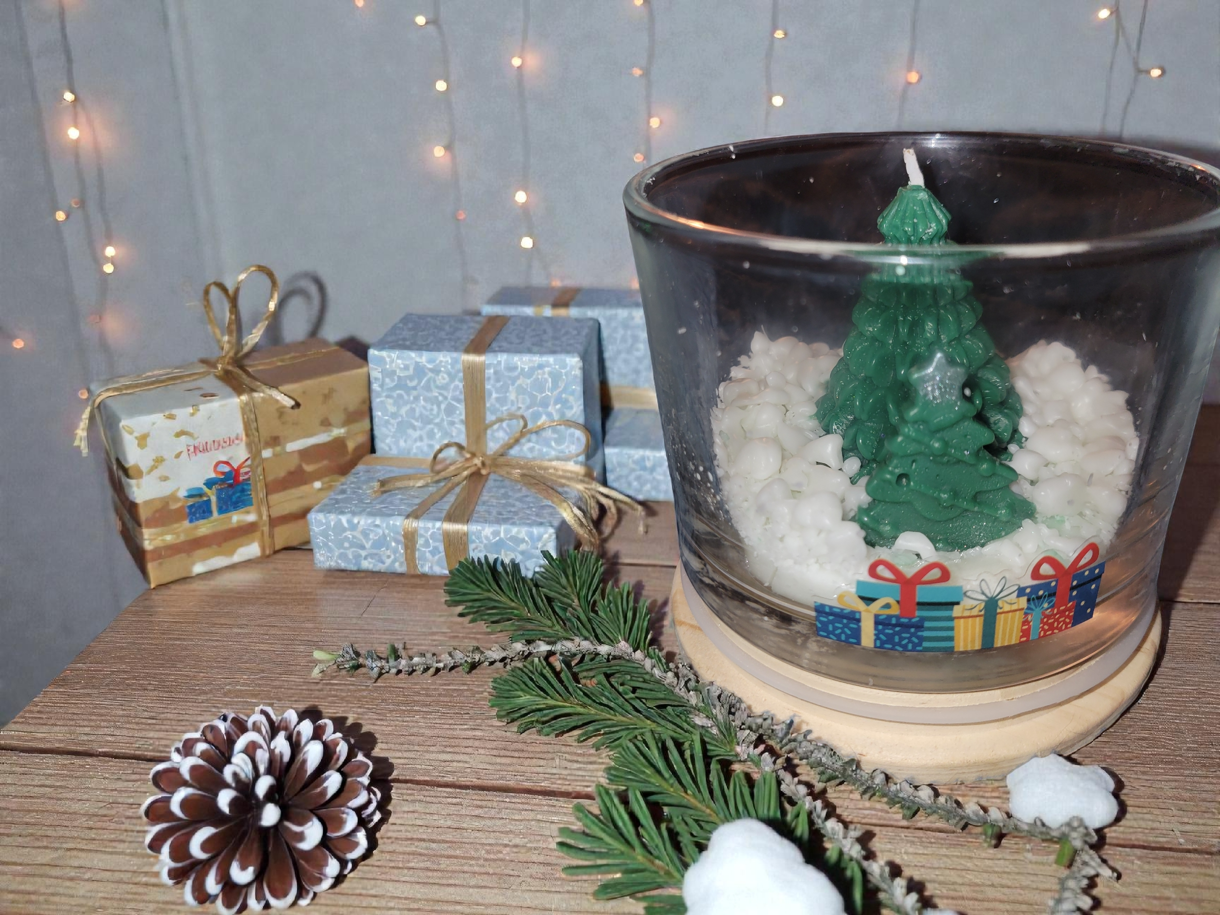 Decorative Christmas scene with a glass bowl containing a green Christmas tree candle surrounded by white decorative balls, along with wrapped gifts, pinecone, and pine branch with snow, on a wooden surface, with blurry string lights in background.