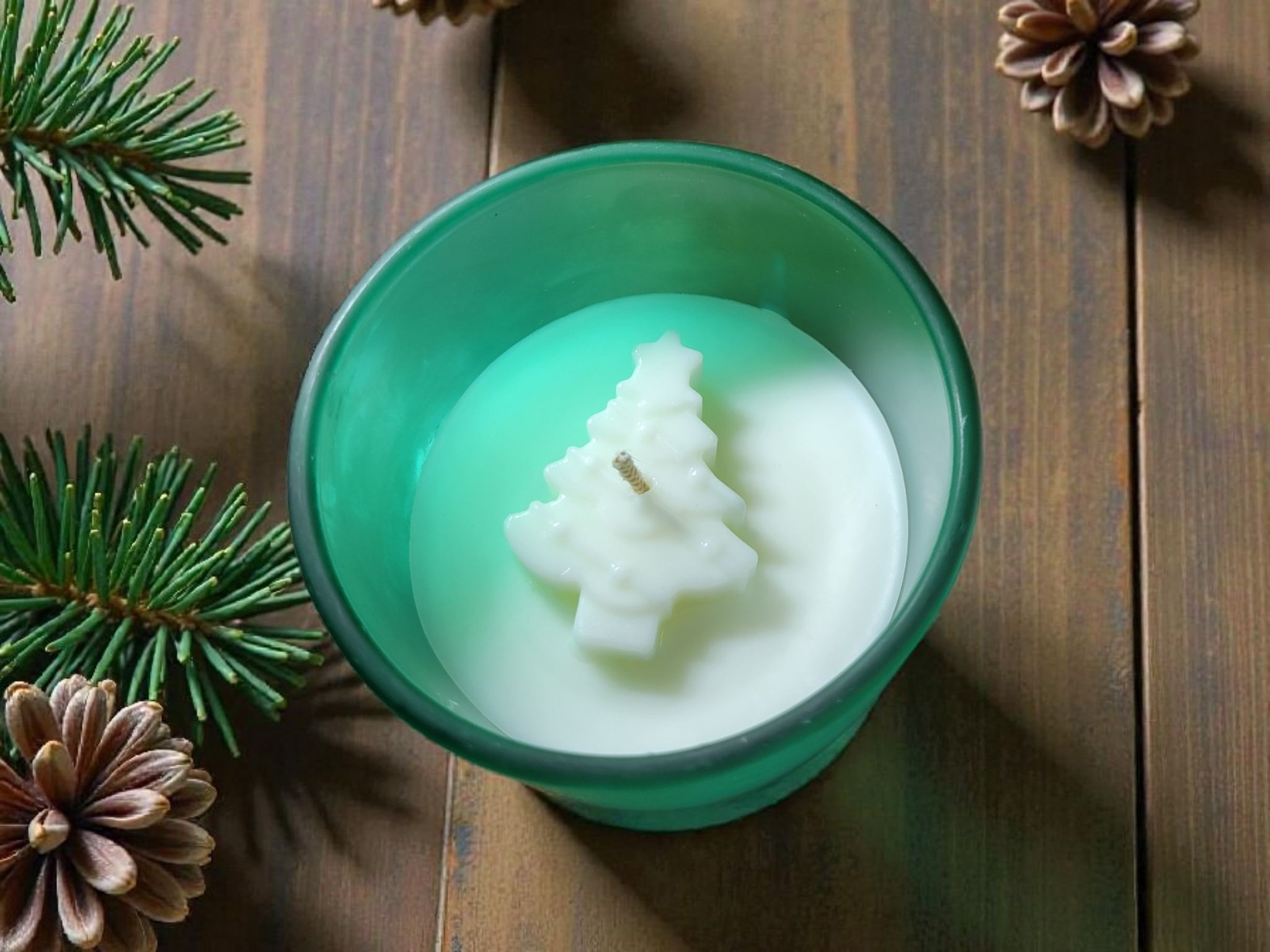 A green glass candle holder with a white candle shaped like a Christmas tree, with a small wick on top, placed on a wooden surface surrounded by pinecones and evergreen branches.