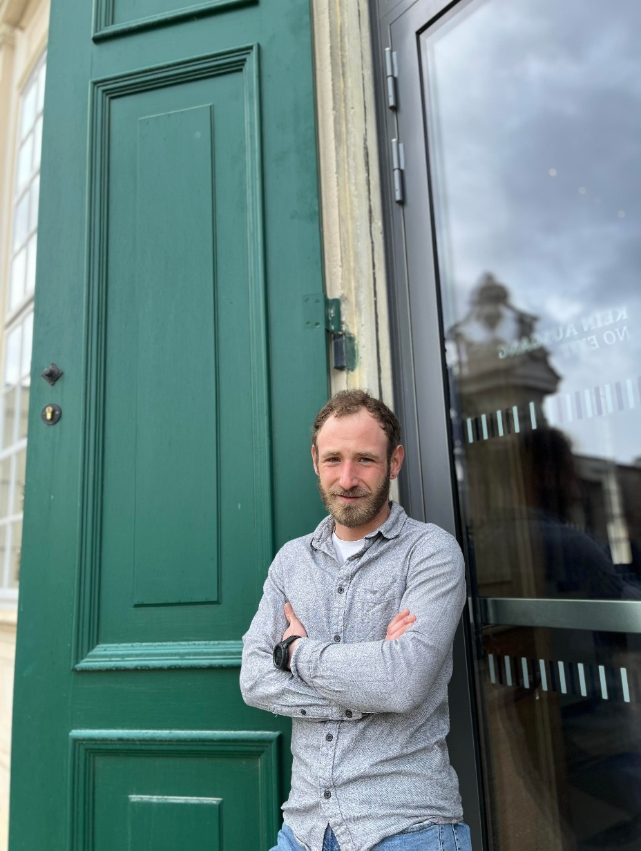 A man with a beard and mustache standing with arms crossed outside a building, in front of a green door and glass window, with a cloudy sky reflected on the glass.