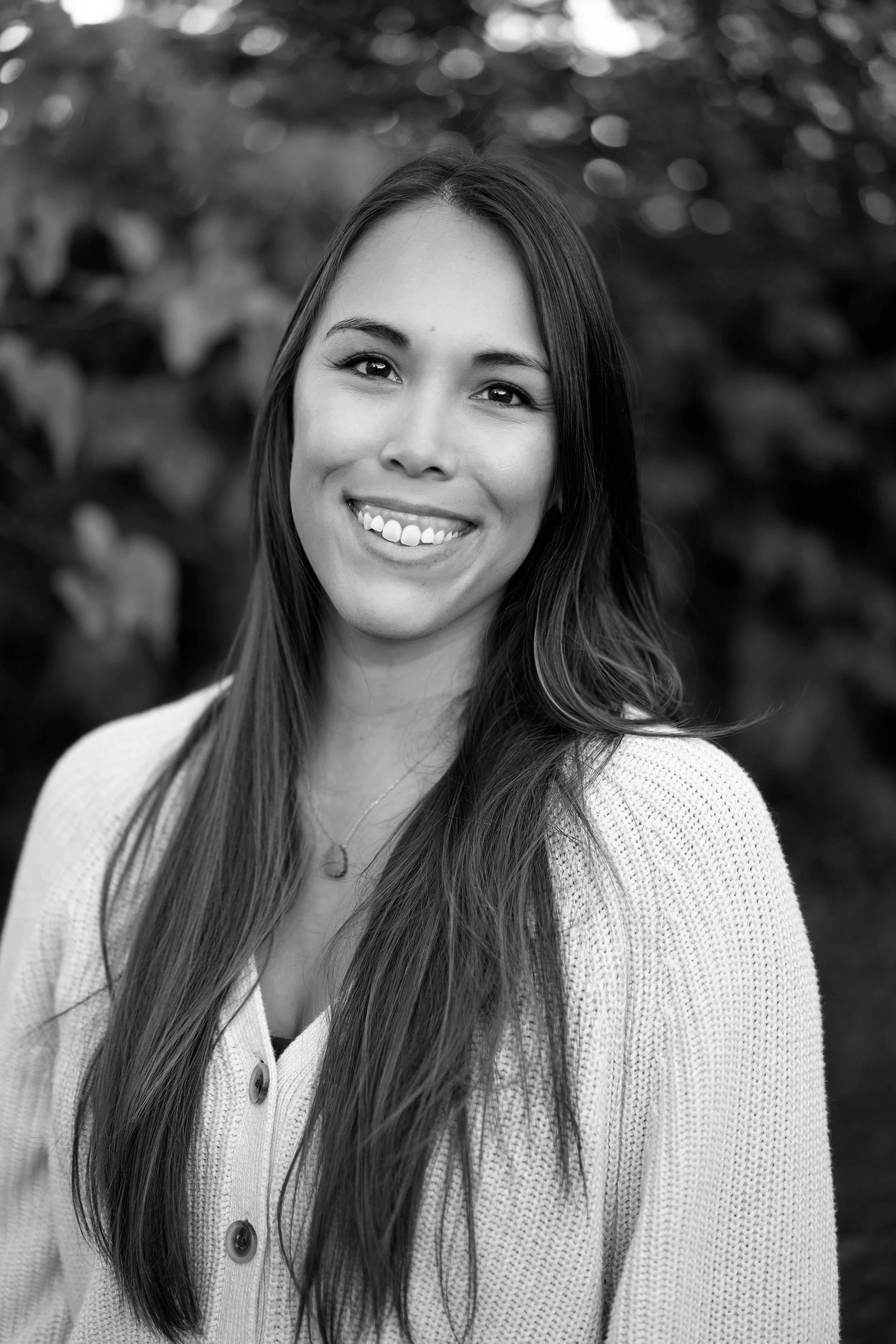 Black and white portrait of a young woman smiling outdoors, wearing a cardigan and necklace.
