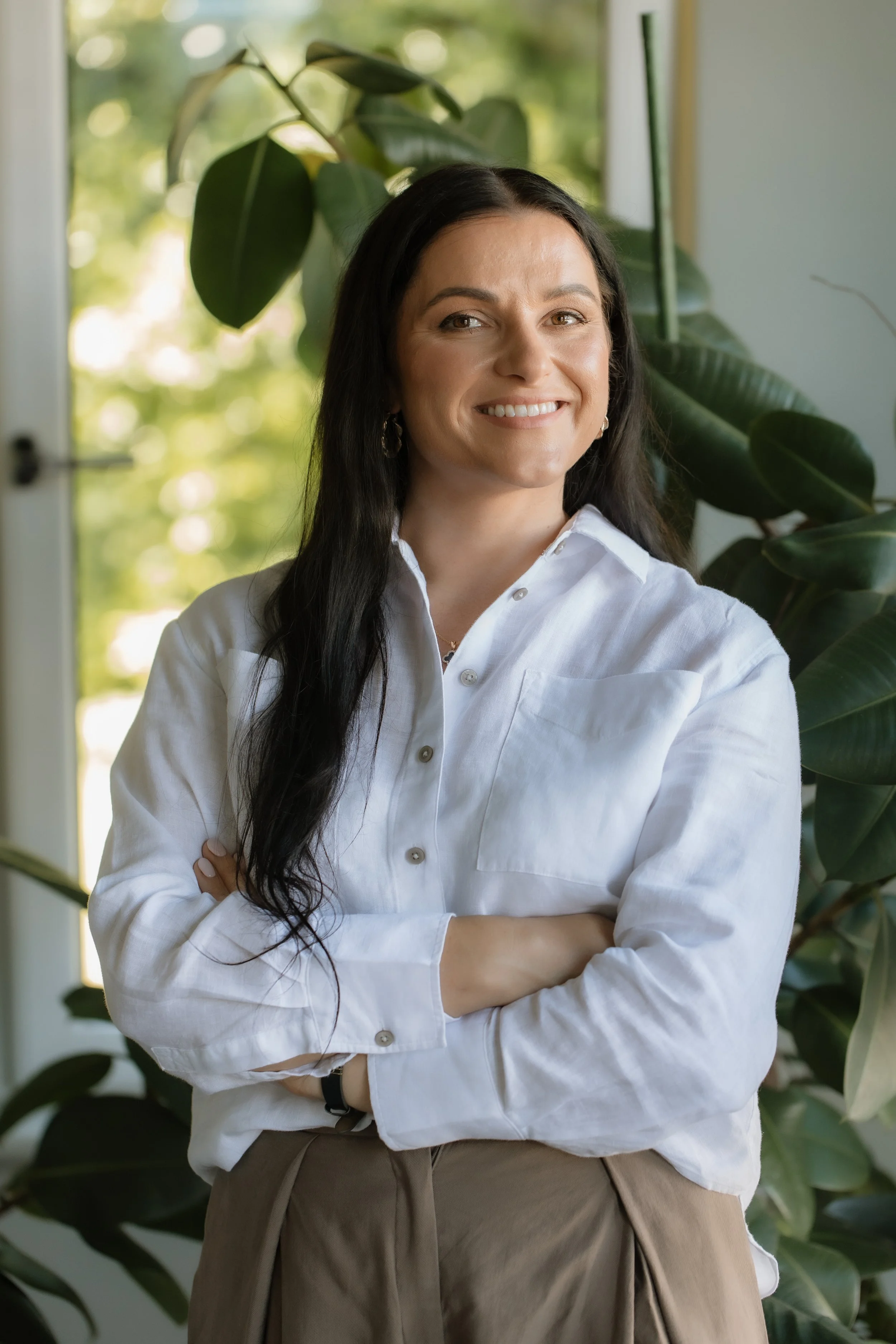 Smiling woman with long dark hair standing with arms crossed in front of large green houseplants, wearing a white button-up shirt and tan pants, with sunlight coming through a window in the background.