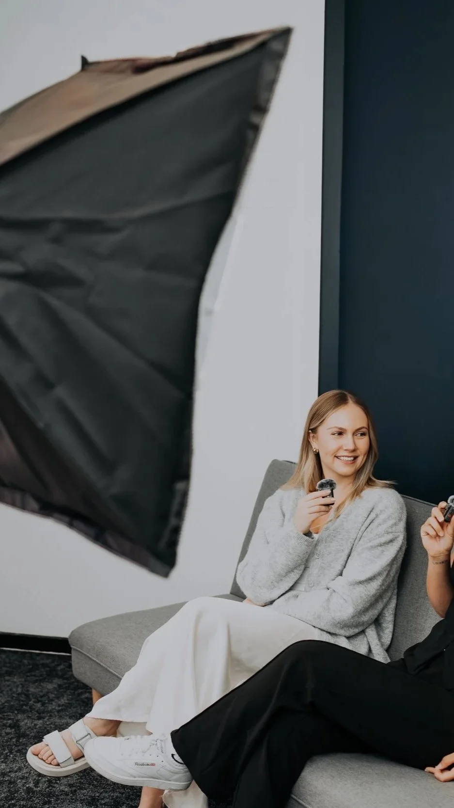 Two women sitting on a gray sofa during a photoshoot, one smiling and holding makeup brushes, with professional lighting equipment visible.
