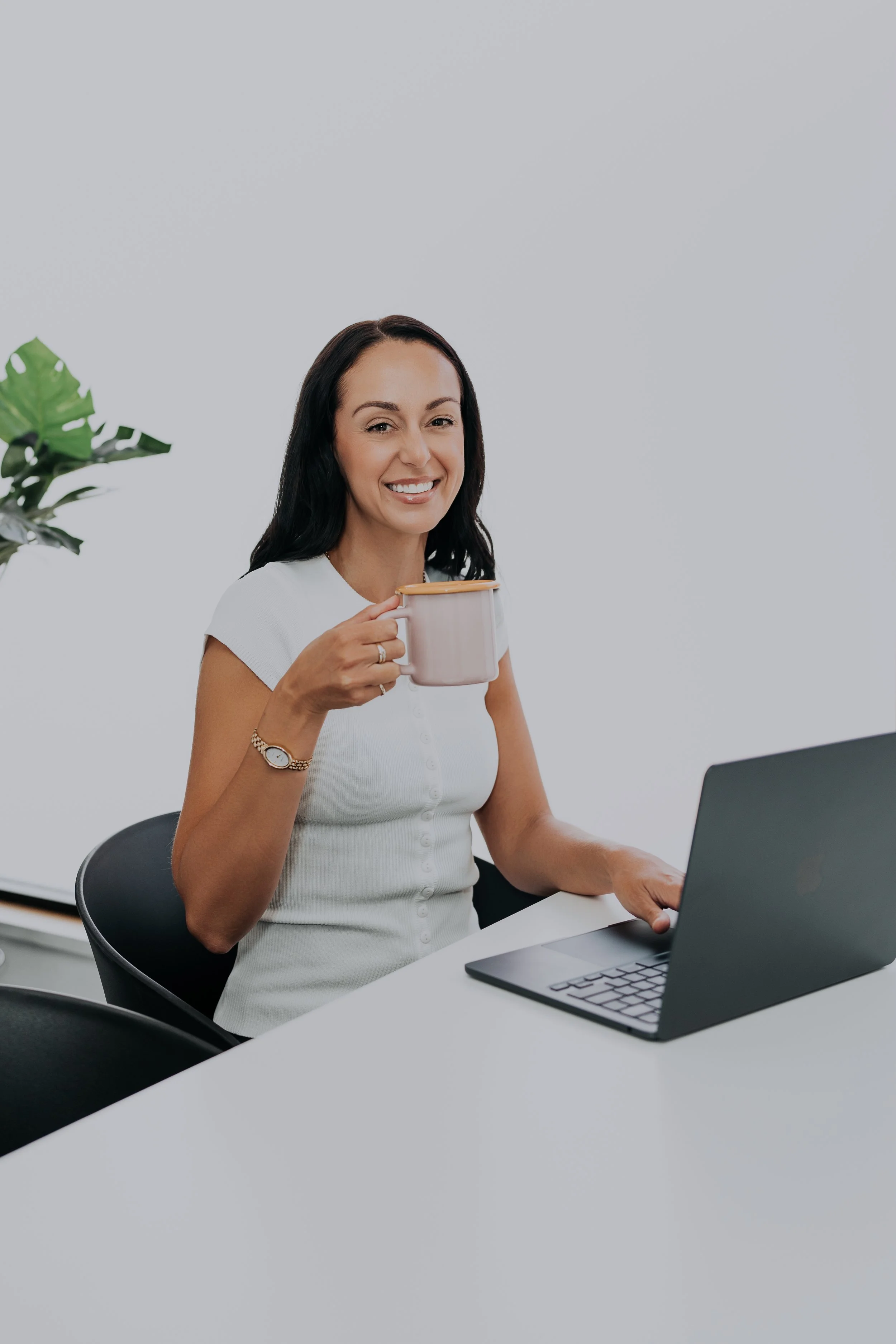 A woman sitting at a white desk, holding a mug, and working on a laptop with a plant nearby.