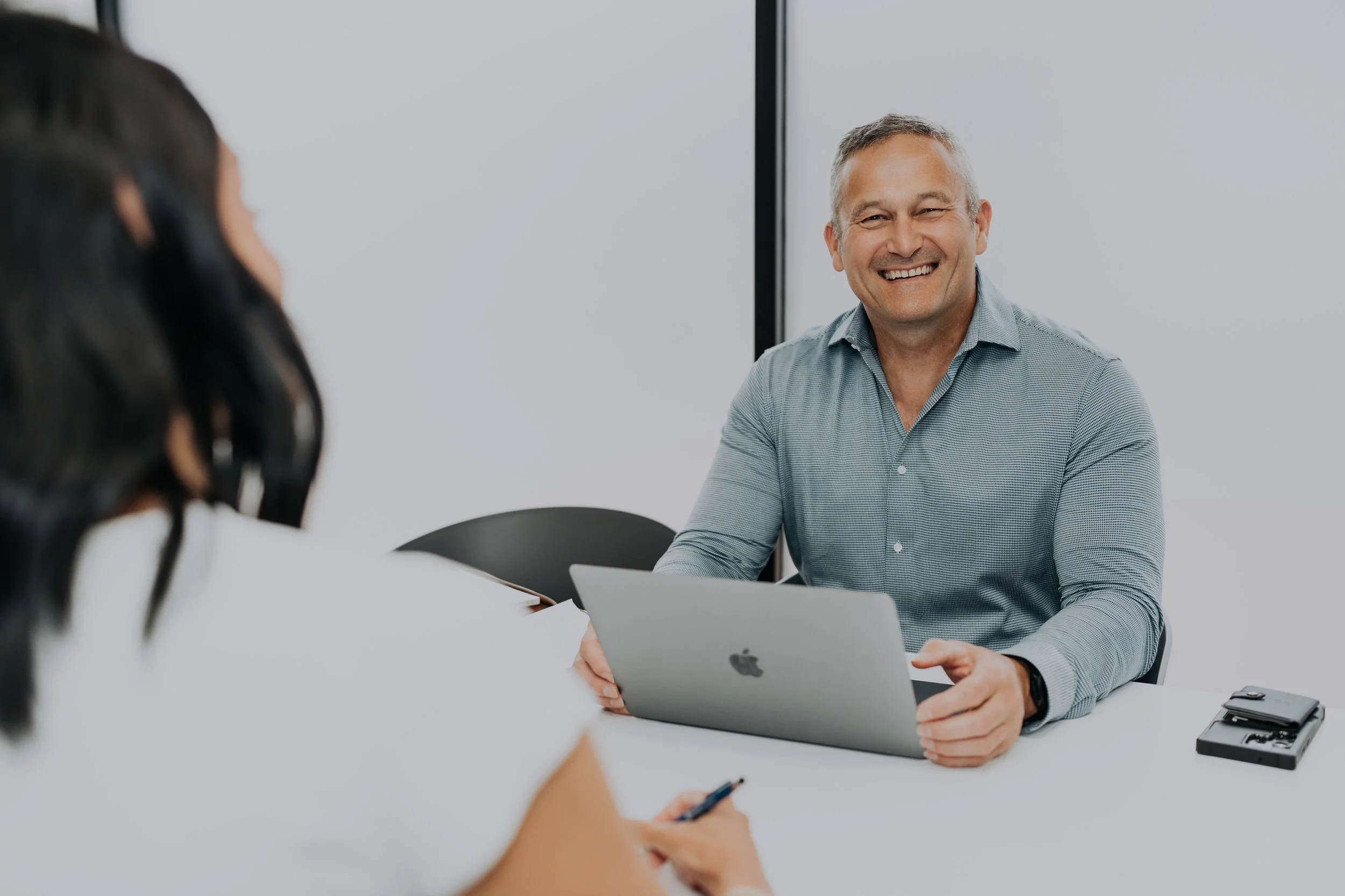 A man smiling and sitting at a table with a laptop, engaging in a conversation with a woman in a business meeting.