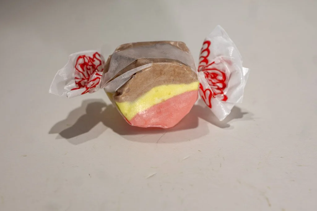 A wrapped piece of banana split saltwater taffy featuring three distinct layers: brown (chocolate), yellow (banana), and pink (strawberry). Twist-wrapped in clear wax paper with red lettering on each end.