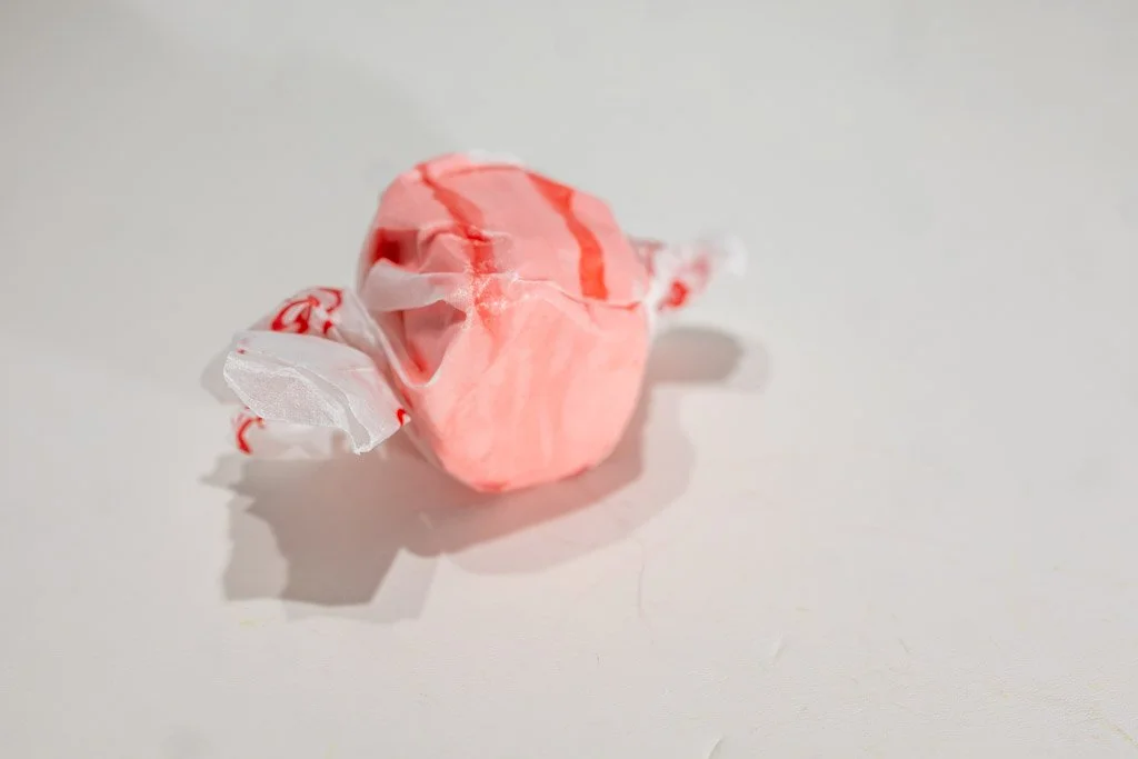 A wrapped piece of strawberry saltwater taffy in a light pink color with soft red swirls, twist-wrapped in clear wax paper with red lettering on each end. A fruity, nostalgic favorite from Sea Bird Gifts & Candy in Westport, WA.