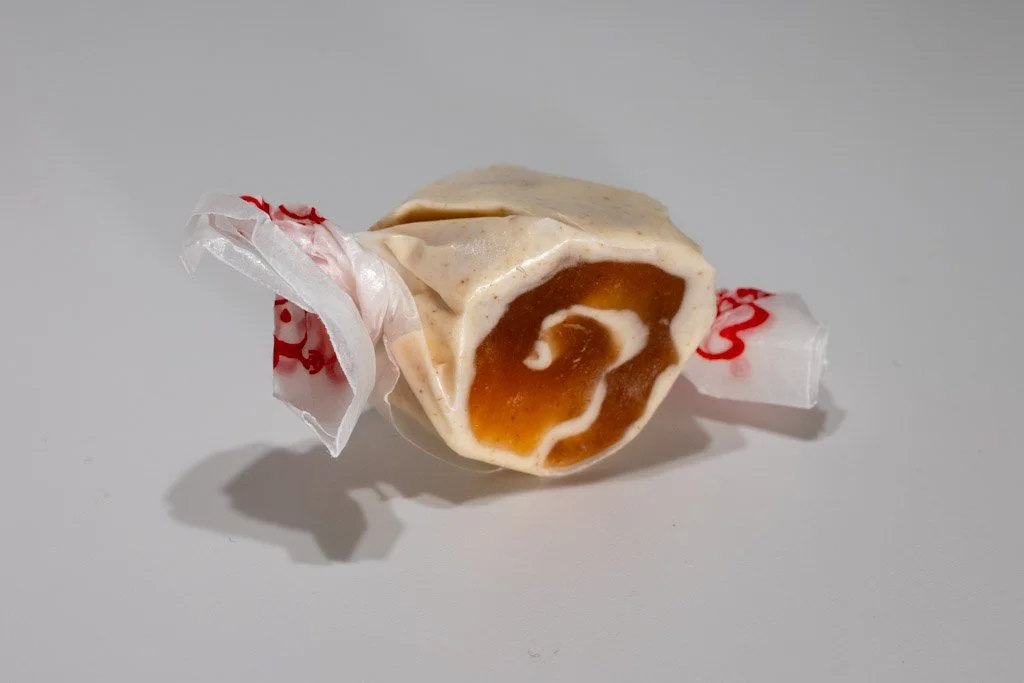 A salt water taffy with a light beige outer layer and a rich brown cinnamon swirl in the center, resembling a cinnamon roll. Wrapped in translucent wax paper with red print.